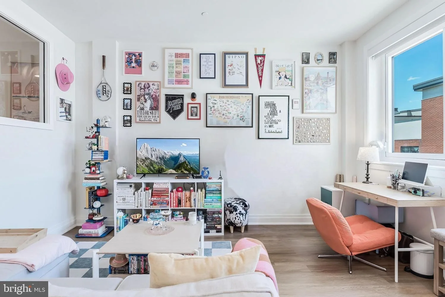 A bright living room with a white wall decorated with framed art and photos, a flat-screen TV on a white bookshelf filled with books, a cozy lounge chair, a small desk with a lamp and computer near a large window showing a blue sky and buildings outs
