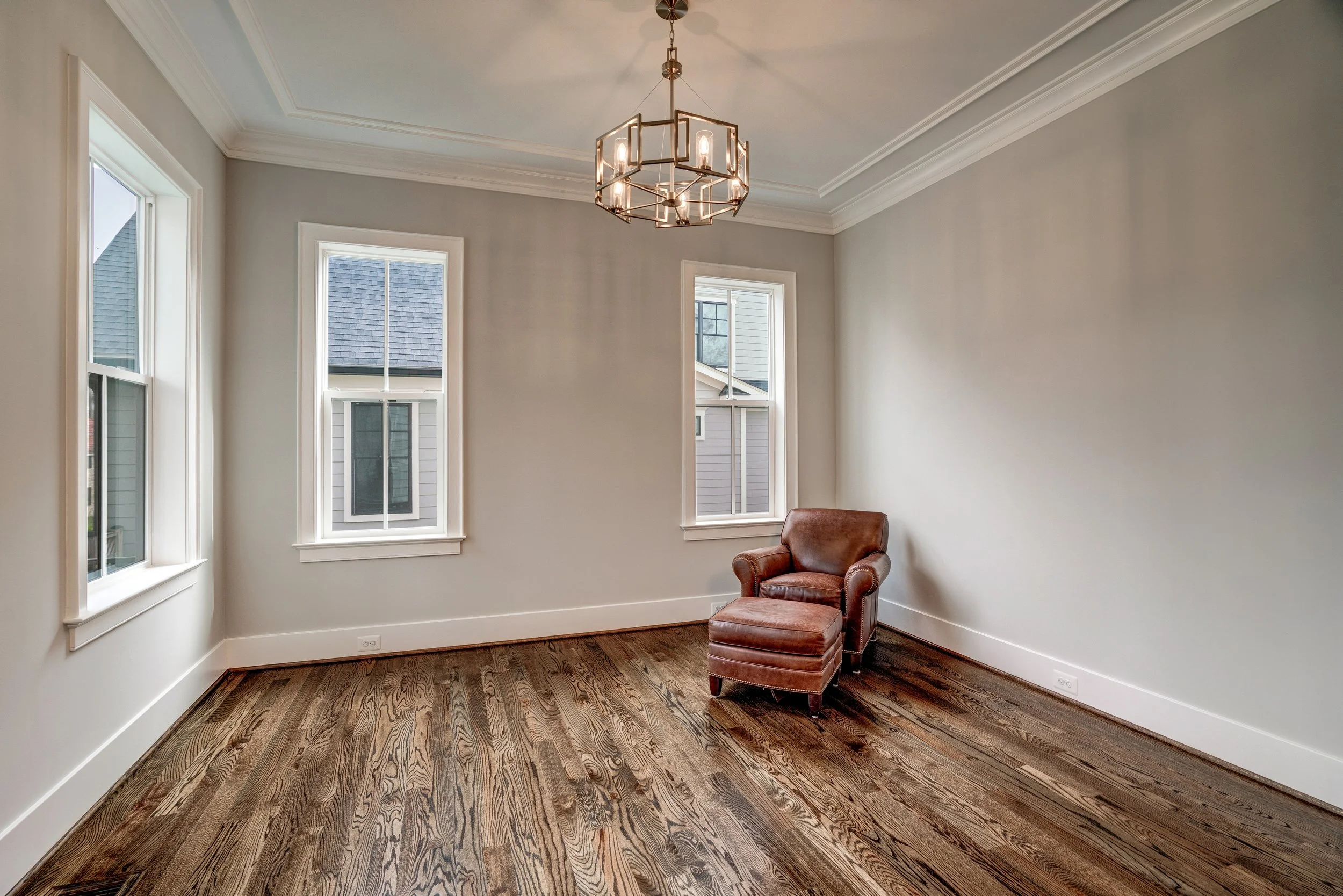 A corner room with three windows, white walls, crown molding, hardwood flooring, a leather armchair, and a modern geometric chandelier.