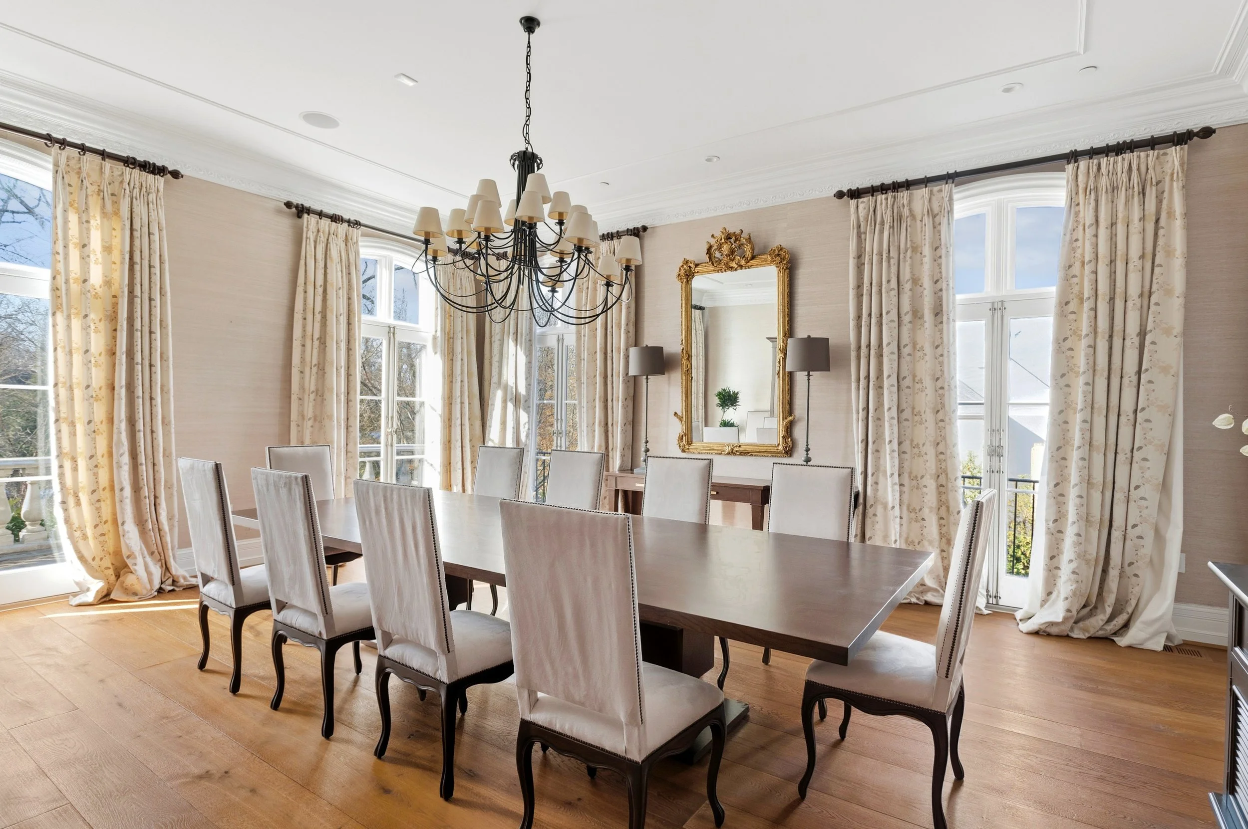 Elegant dining room with large windows, cream-colored curtains, a rustic wooden table, white upholstered chairs with dark legs, a gold ornate mirror, two grey floor lamps, a black chandelier with white shades, and a small potted plant on a side table