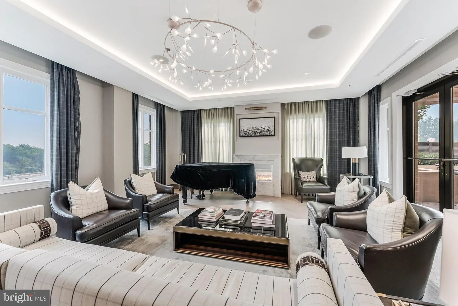 Elegant living room with a grand piano, six armchairs with throw pillows, a central coffee table with books, a fireplace, large windows with sheer curtains, and a modern chandelier with branch-like light fixtures.