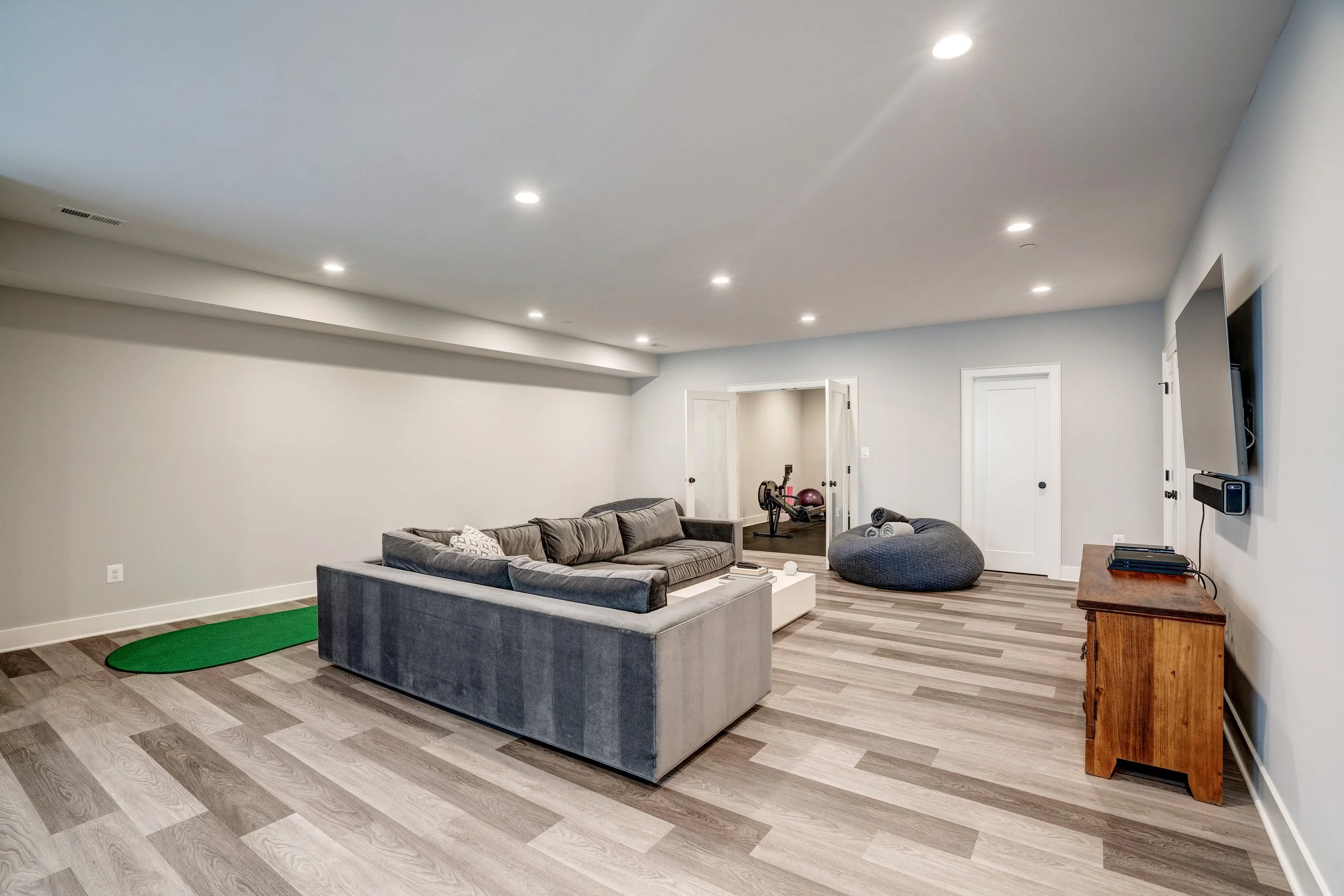 Living room with gray sectional sofa, workout equipment in a room behind, wood flooring, wall-mounted TV, and a wooden cabinet.