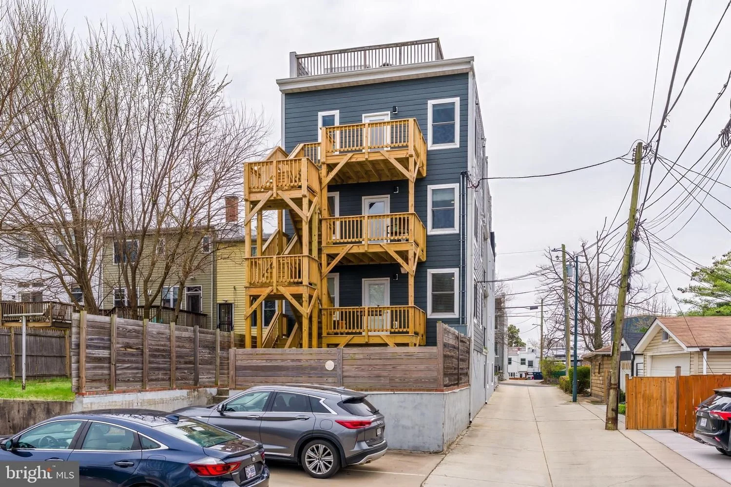 Four-story blue residential building with multiple wooden balconies, situated in an urban neighborhood with parked cars, trees, and overhead power lines.