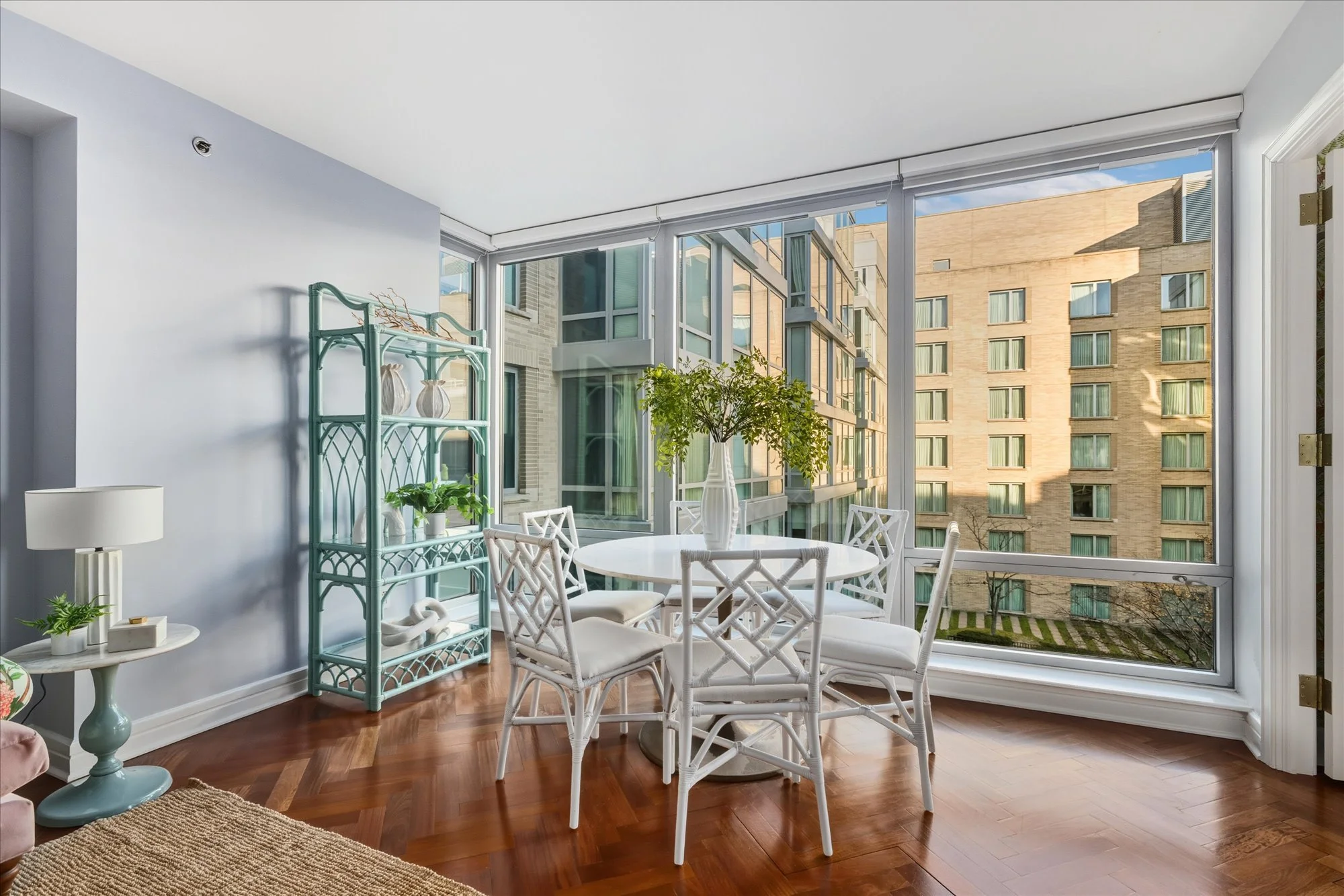 Bright sunlit interior of a dining area with a round white table and white chairs, large windows showing neighboring apartment buildings, potted plants, and a small teal shelving unit against a light blue wall.