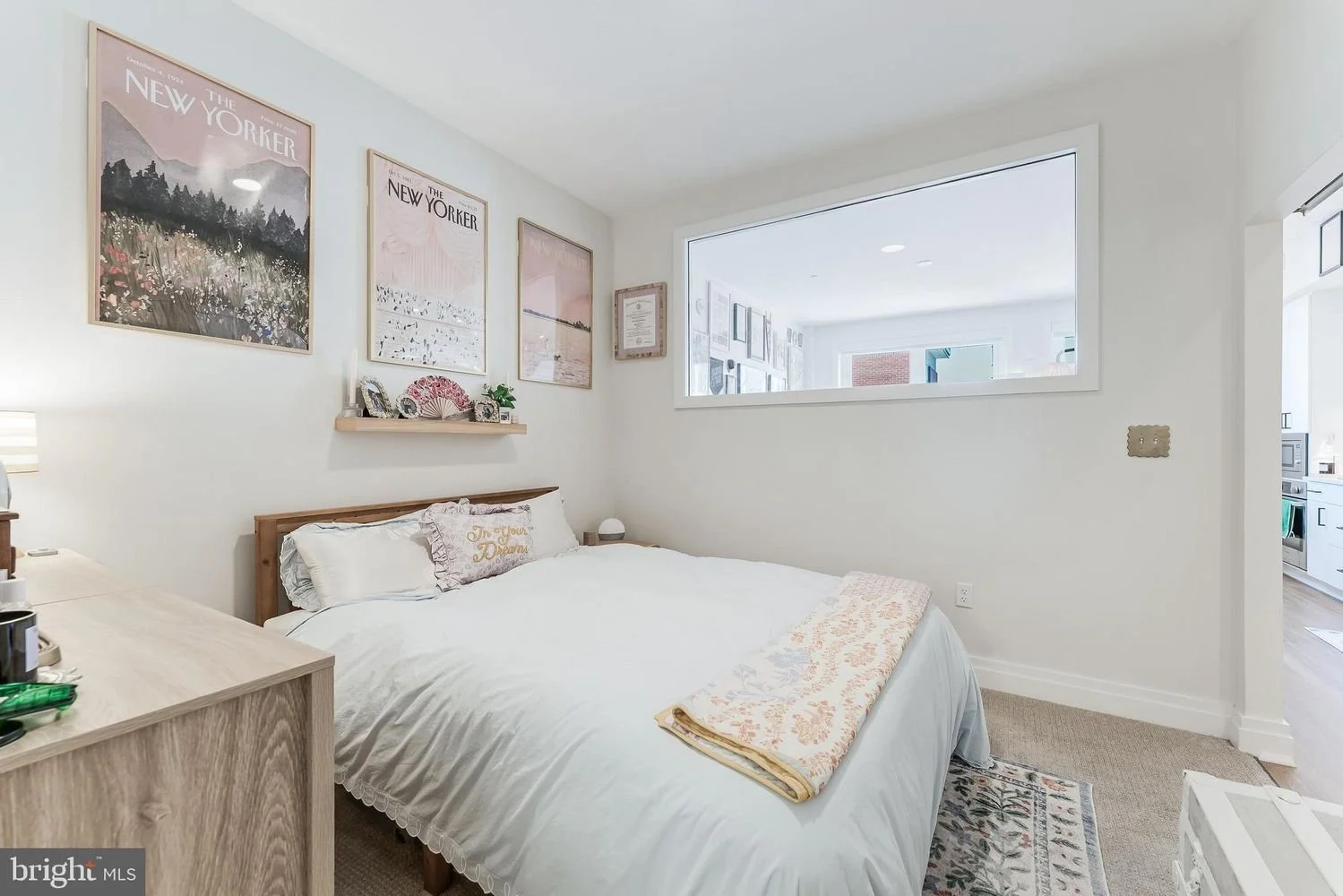 Clean, bright bedroom with framed art on white walls, a bed with white bedding and decorative pillows, and a window looking into another room with art on the walls.