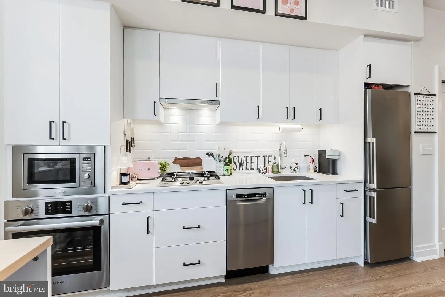 White kitchen with stainless steel appliances, white cabinets, subway tile backsplash, and wood flooring. Items on the counter include a pink toaster, a jar, a soap dispenser, and a black coffee machine. A sign reads "SWEET TEA" behind the stove.