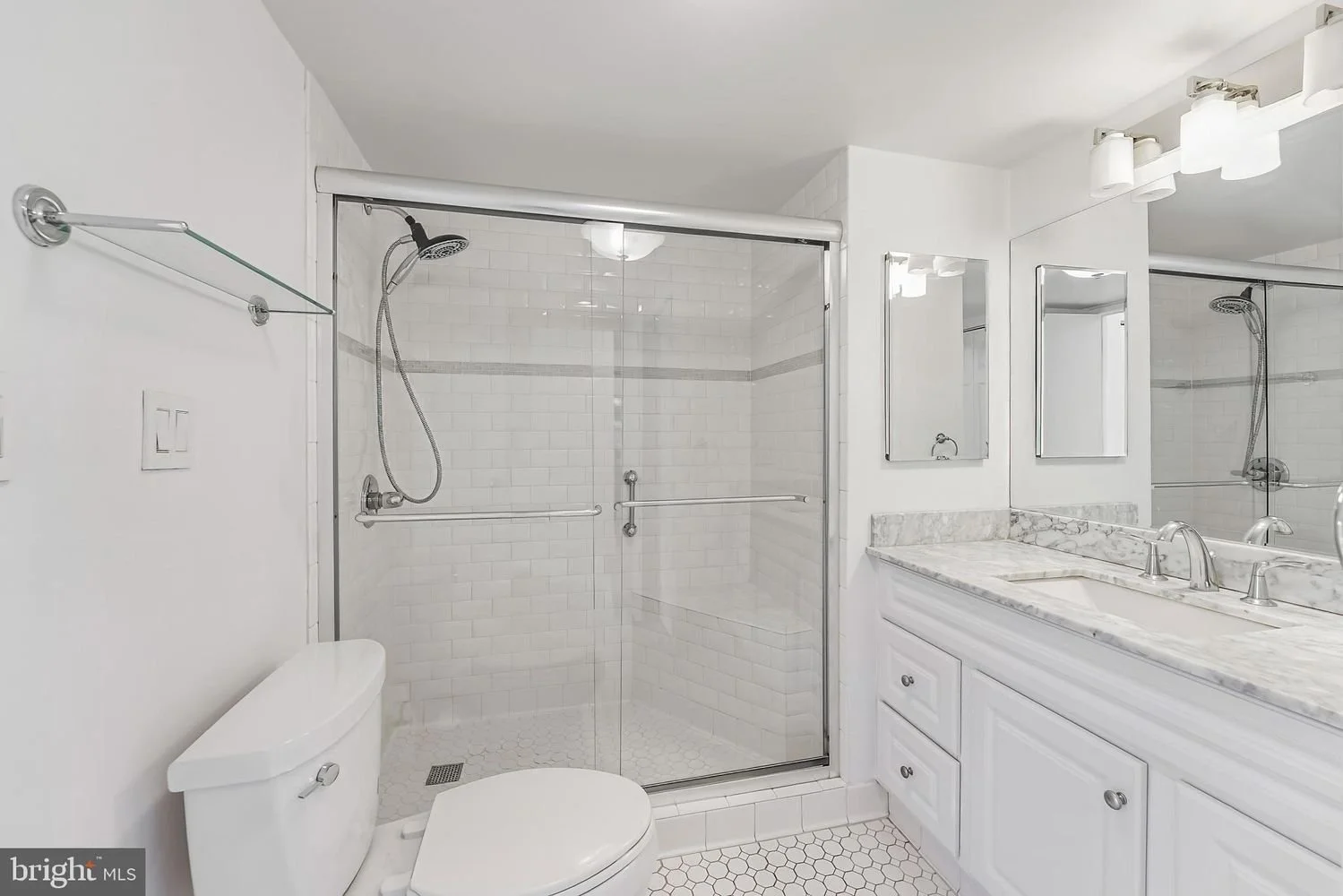 Bathroom with glass shower enclosure, white tiled walls, white vanity with marble countertop, mirror, and lighting fixtures.