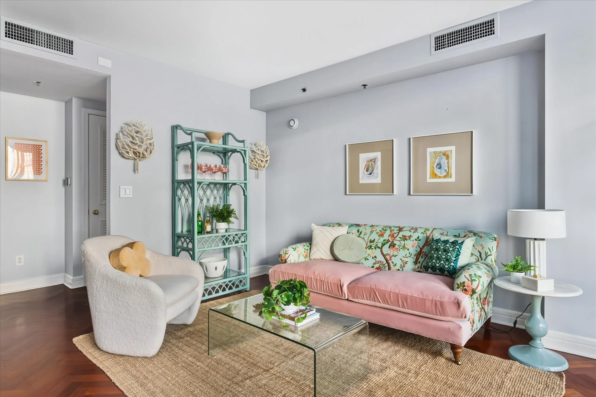 A living room with a floral sofa, a white armchair, a glass coffee table with a plant and books, a patterned area rug, framed artwork, and decorative wall pieces, with light-colored walls and hardwood floors.