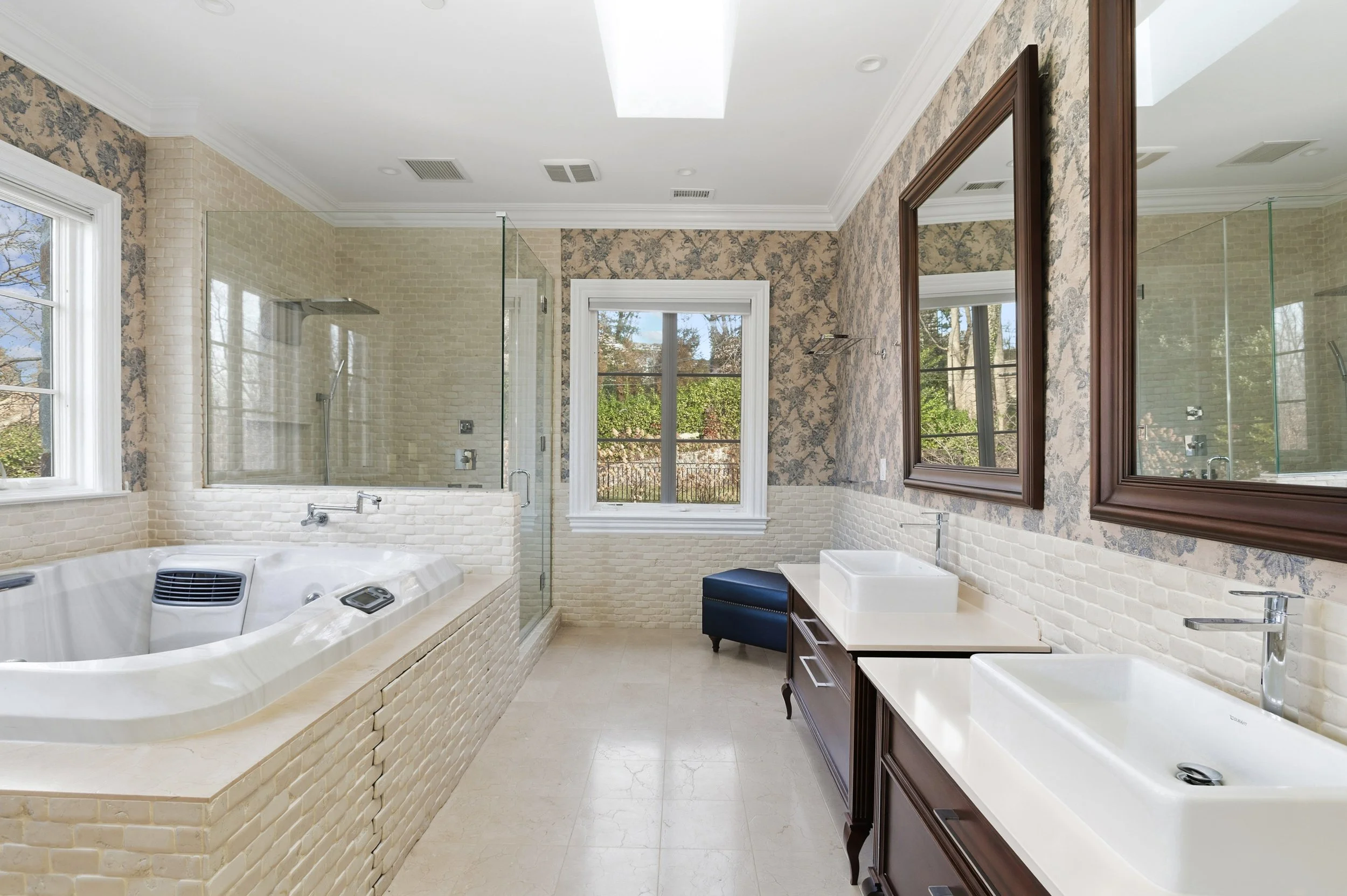 Bathroom with a jacuzzi tub, a glass-enclosed shower, a window, mirror, and vanity with dual sinks, finished with a mix of floral and brick wall decor.