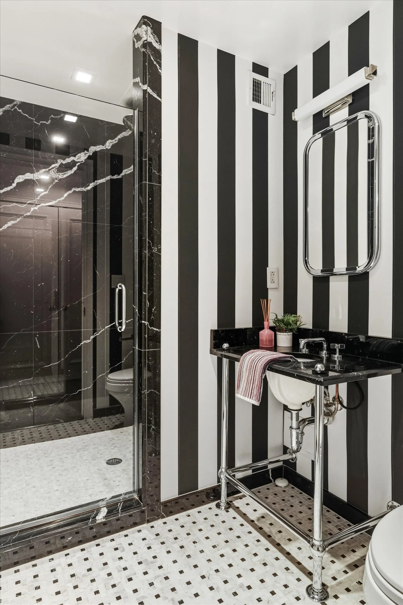 A modern bathroom with black and white striped walls, a black marble shower with a glass door, a black marble sink counter, a white porcelain sink, a mirror with a metal frame, and a small potted plant. The floor has white and black mosaic tiles.
