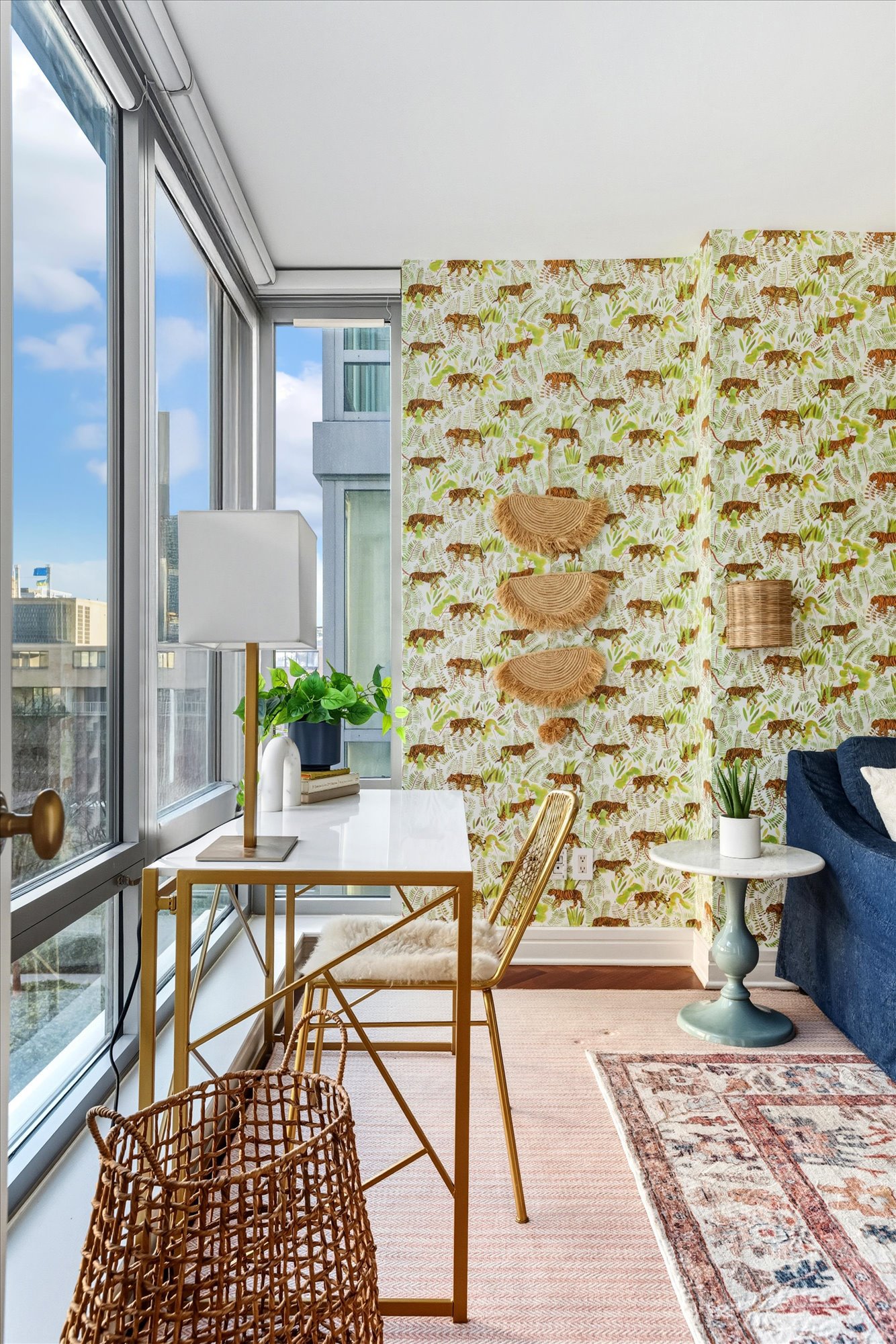 A bright living room with large windows, a white desk with gold accents and a lamp, green plants, a rattan basket, a blue sofa, a small round white table with a potted plant, a decorative rug, and a tropical animal print partition wall.
