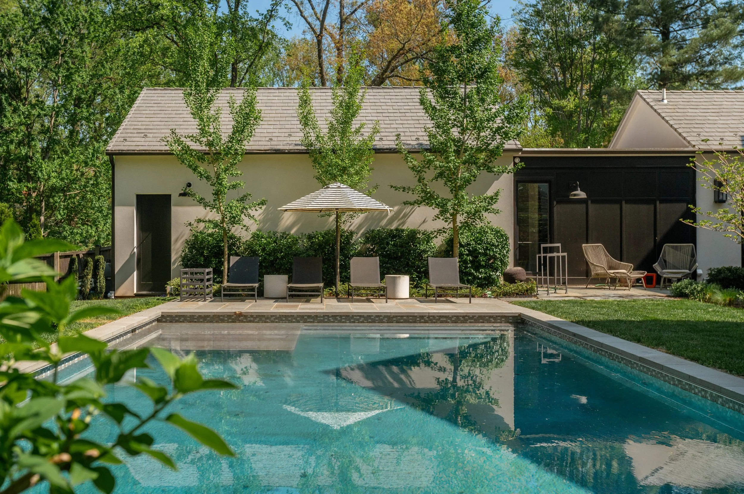 Backyard with a swimming pool, patio seating area, trees, and a modern house in the background.