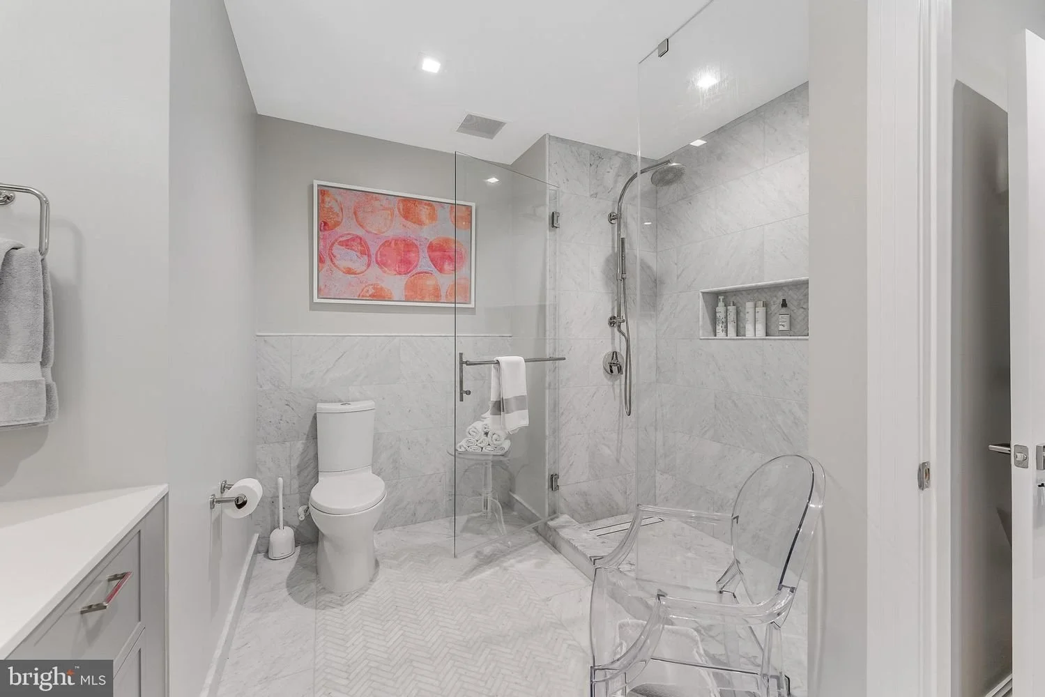 A modern bathroom with a walk-in glass shower, a toilet, and a vanity with a towel rack. The shower has a built-in shelf with toiletries and a textured floor mat. There is a piece of colorful abstract art on the wall above the toilet.
