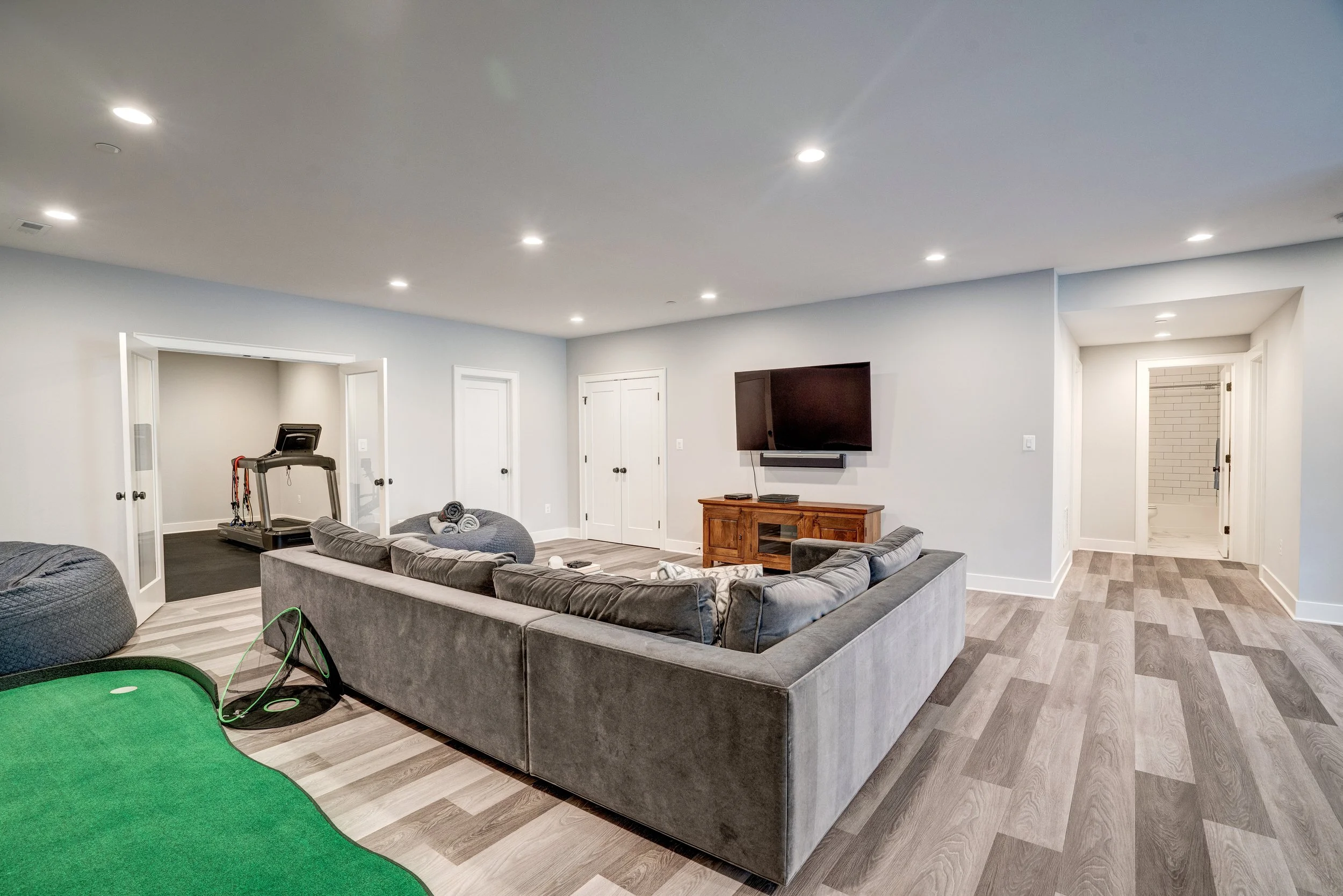 A spacious living room with a gray sectional sofa facing a flat-screen TV mounted on the wall. The room features a wood floor with a herringbone pattern, white walls, and multiple ceiling lights. There is a treadmill in an adjoining room visible thro