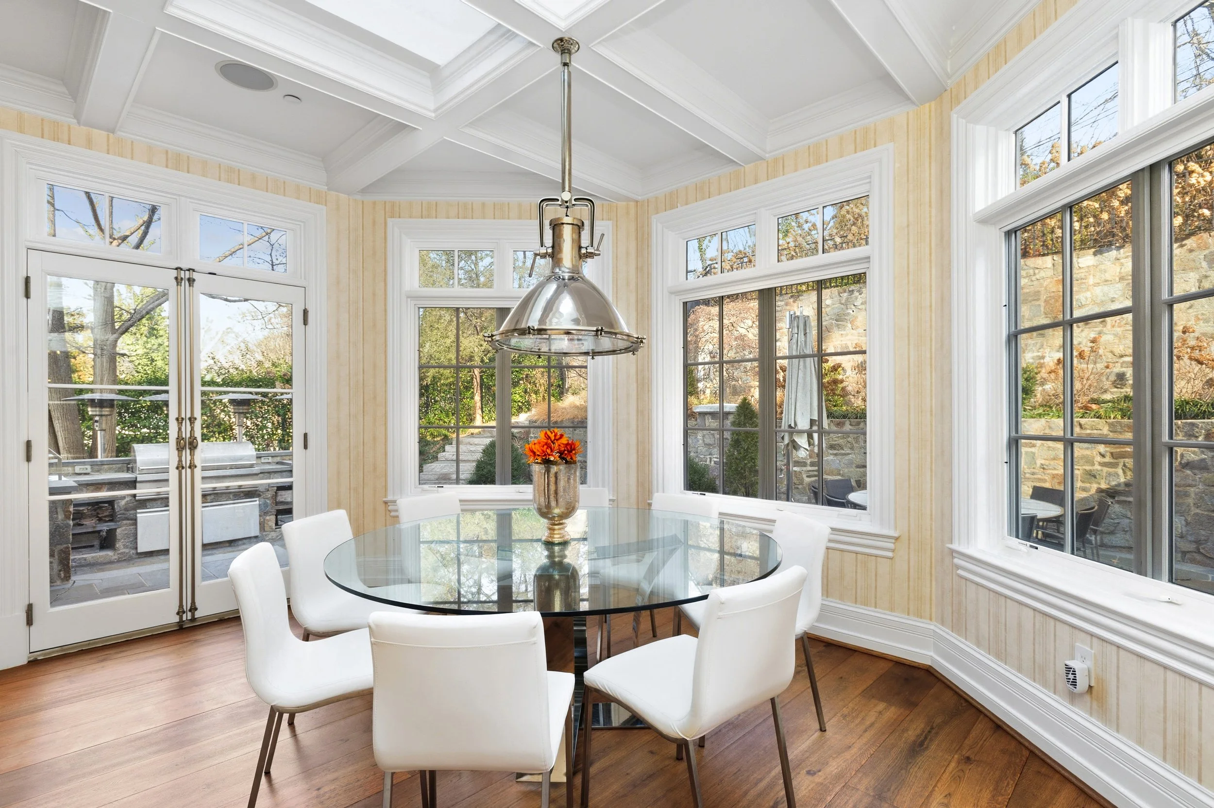 A dining room with large windows and glass doors, a round glass table with white chairs, a metallic pendant light, a floral centerpiece, hardwood floors, and an outdoor patio with a barbecue grill and trees outside.