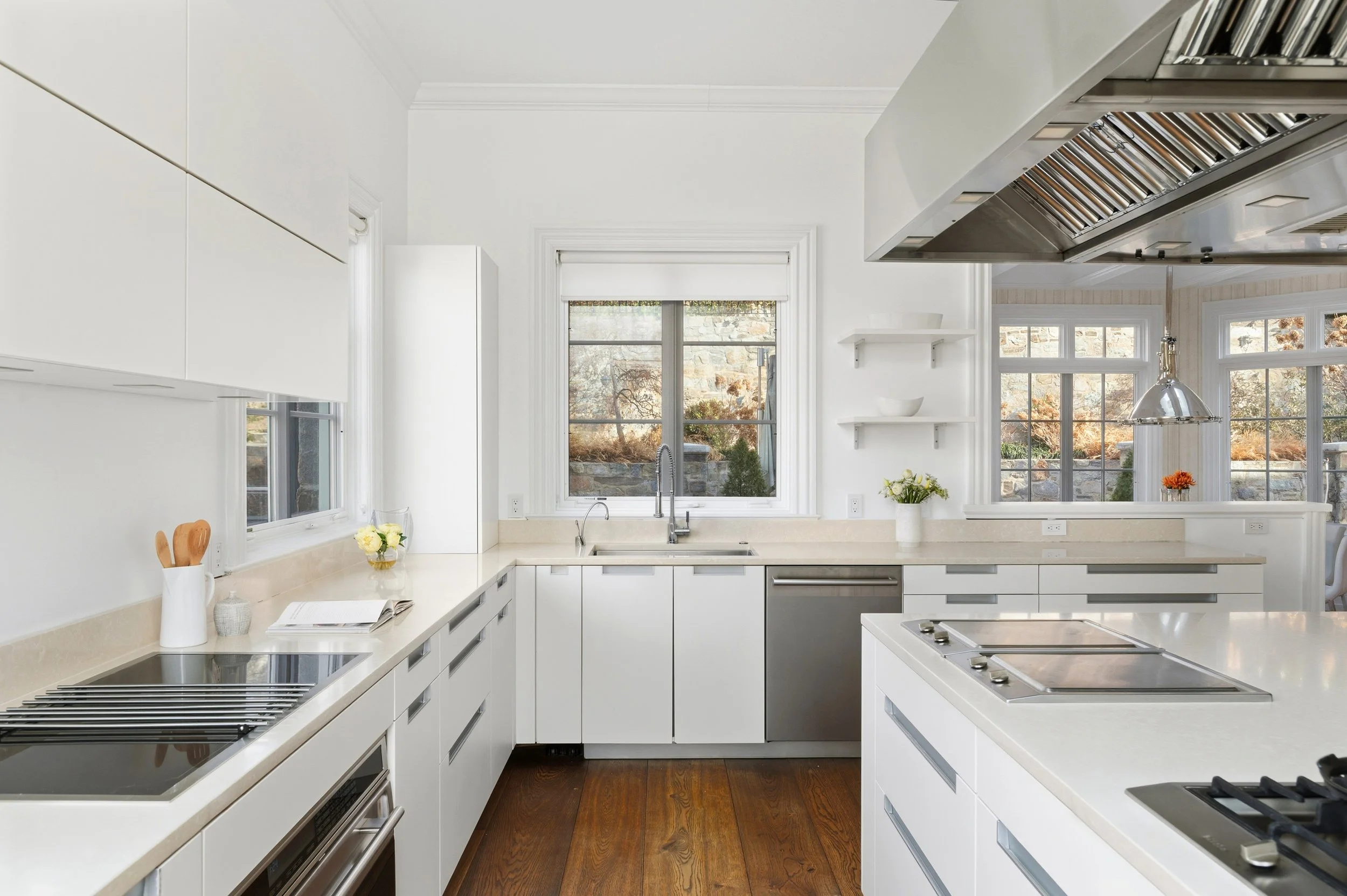 A modern white kitchen with large windows, beige countertops, and wooden flooring. It features stainless steel appliances and open shelves with bowls and decorative items, with natural light coming in.