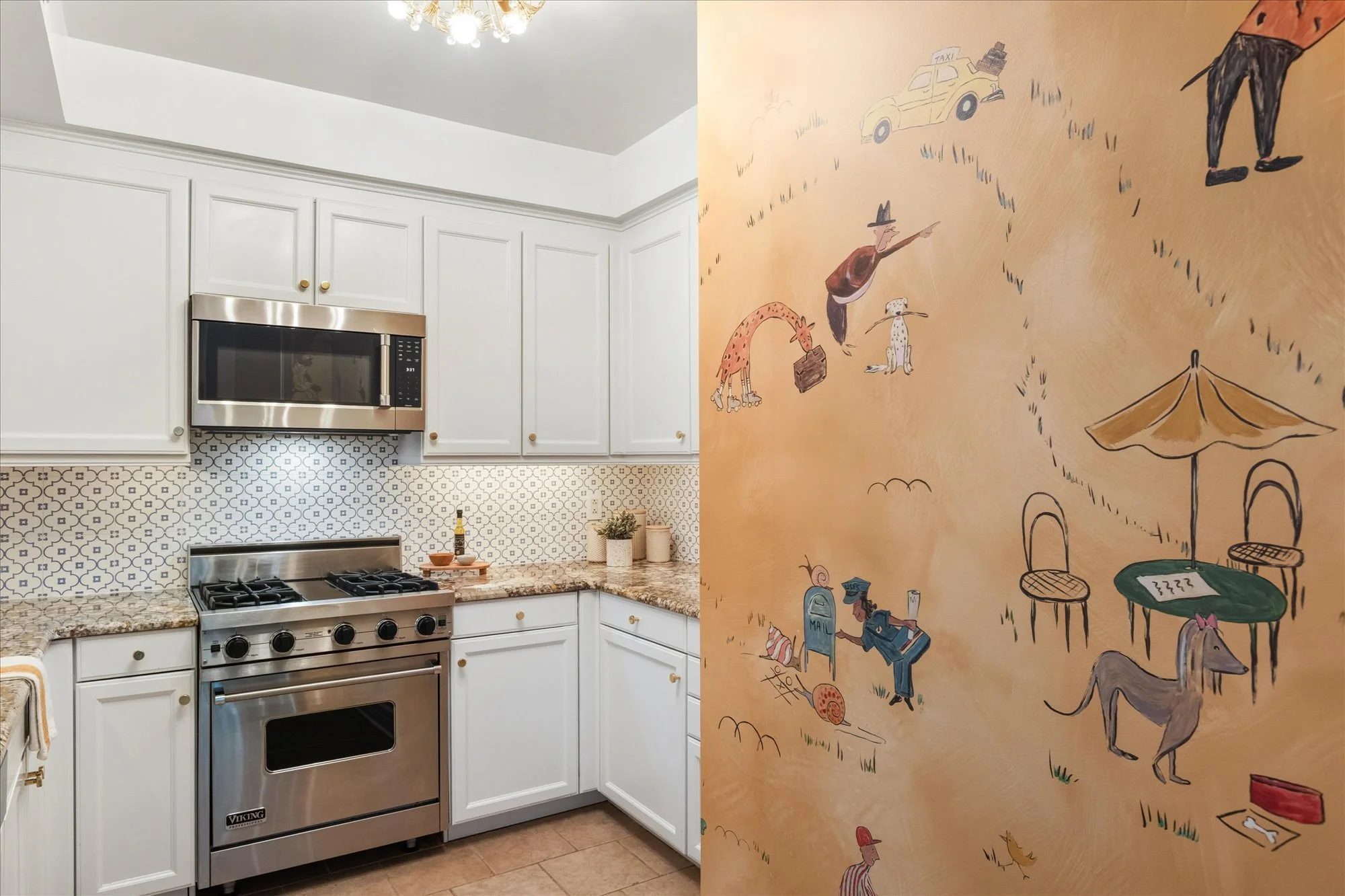 A kitchen with white cabinets, a stainless steel stove and microwave, patterned backsplash, and granite countertops on the left. The right side shows a colorful mural with whimsical drawings of people, animals, and objects on a yellow background.