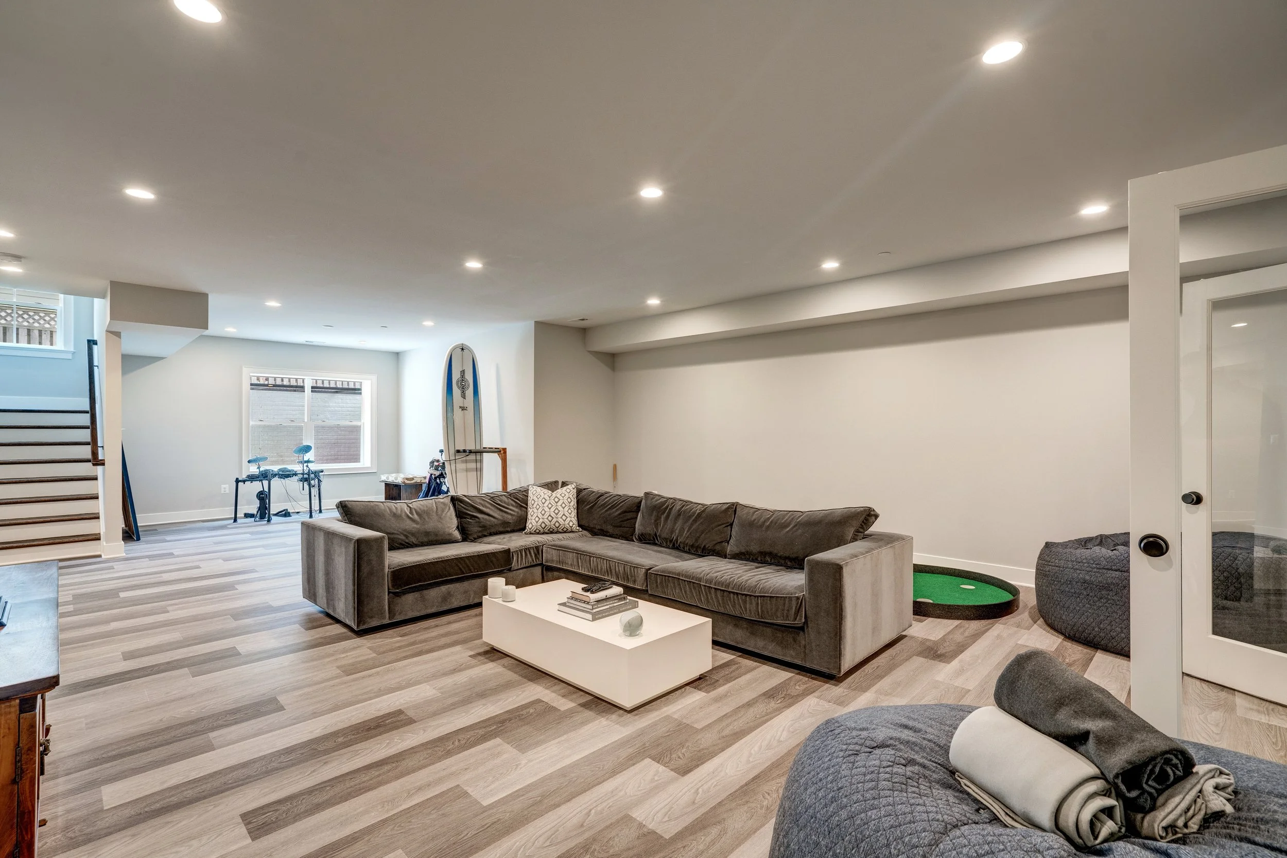 A spacious basement living area with a gray sectional sofa and a white coffee table. The room has a wood-flooring, a window with daylight, and a green mini golf putting hole near a white door. There are several ceiling lights and a power rack with a 
