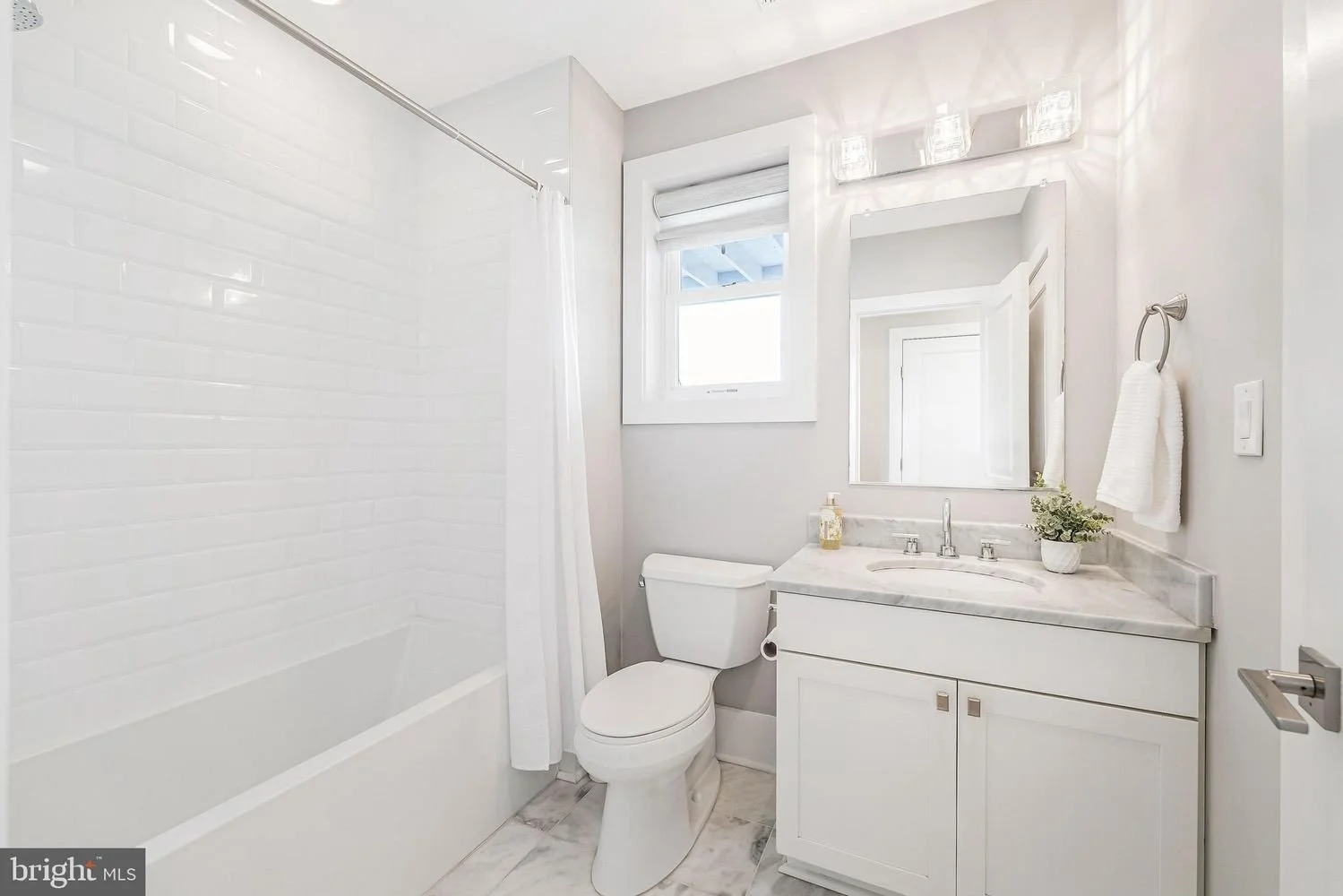 A modern bathroom with a white bathtub, a toilet, and a white vanity with a marble countertop. There's a small window above the toilet, a mirror above the sink, and a towel ring with a white towel on the wall. The bathroom has light gray walls, a mar