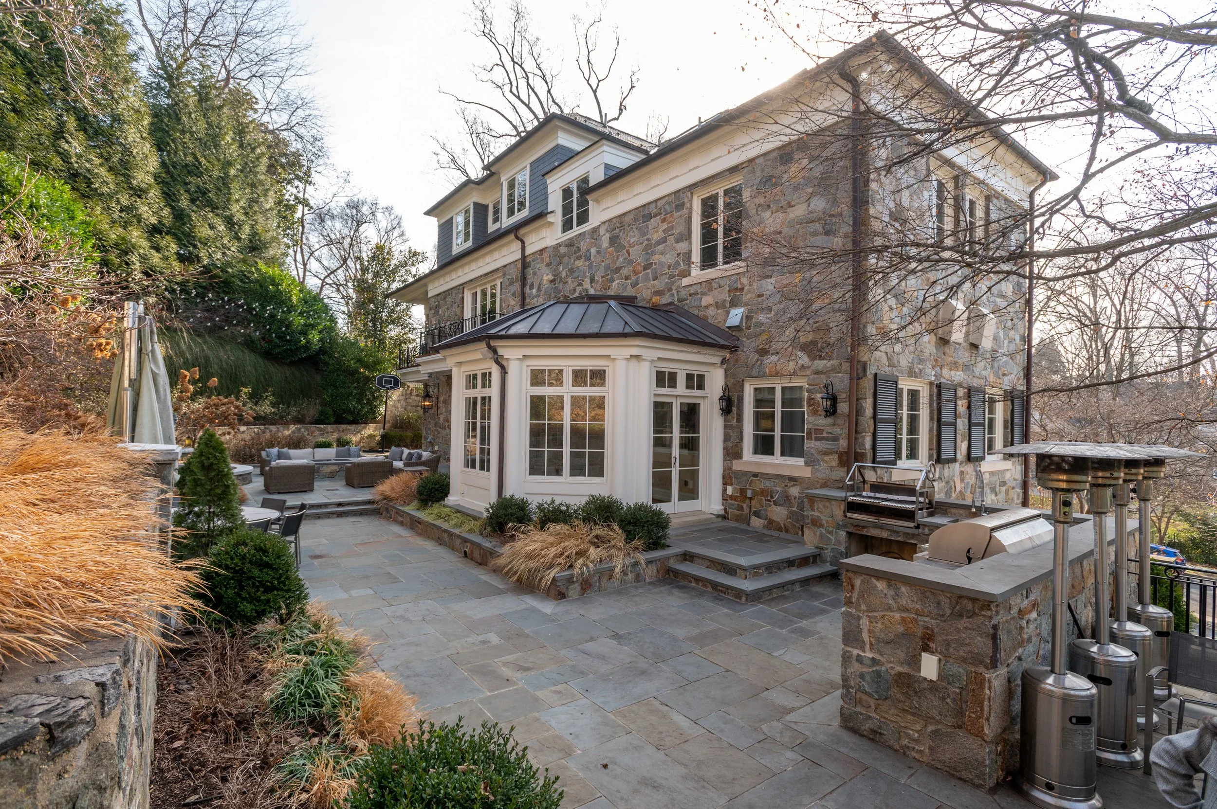 A backyard patio with a stone house, outdoor furniture, a basketball hoop, and trees with no leaves.