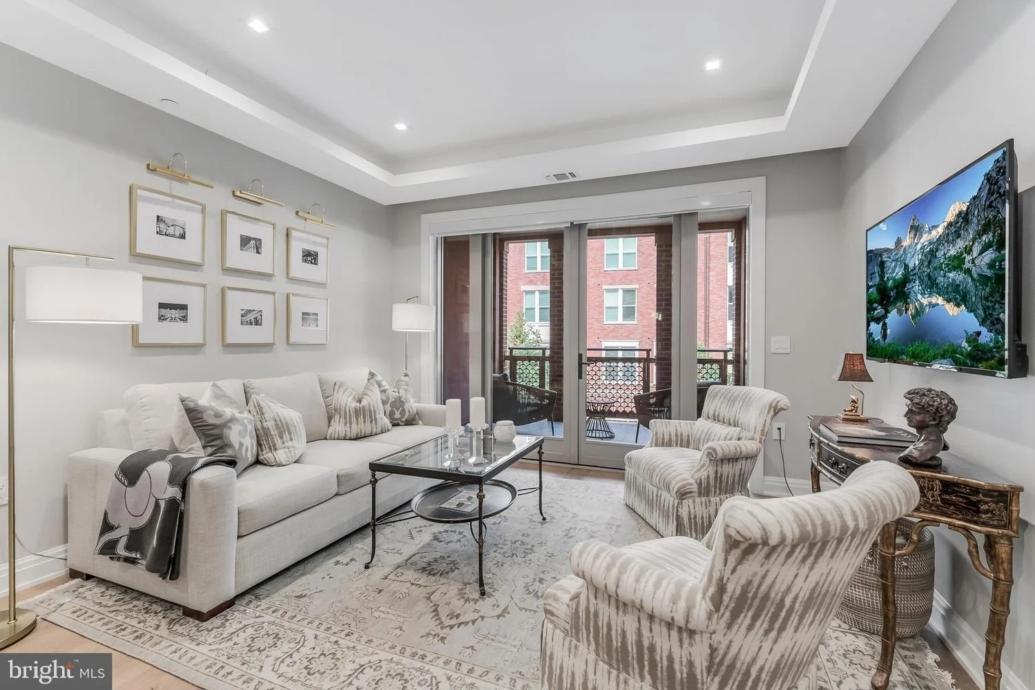 Living room with white sofa, striped armchairs, glass coffee table, wall-mounted TV, sliding glass door leading to balcony, artwork and lamps.
