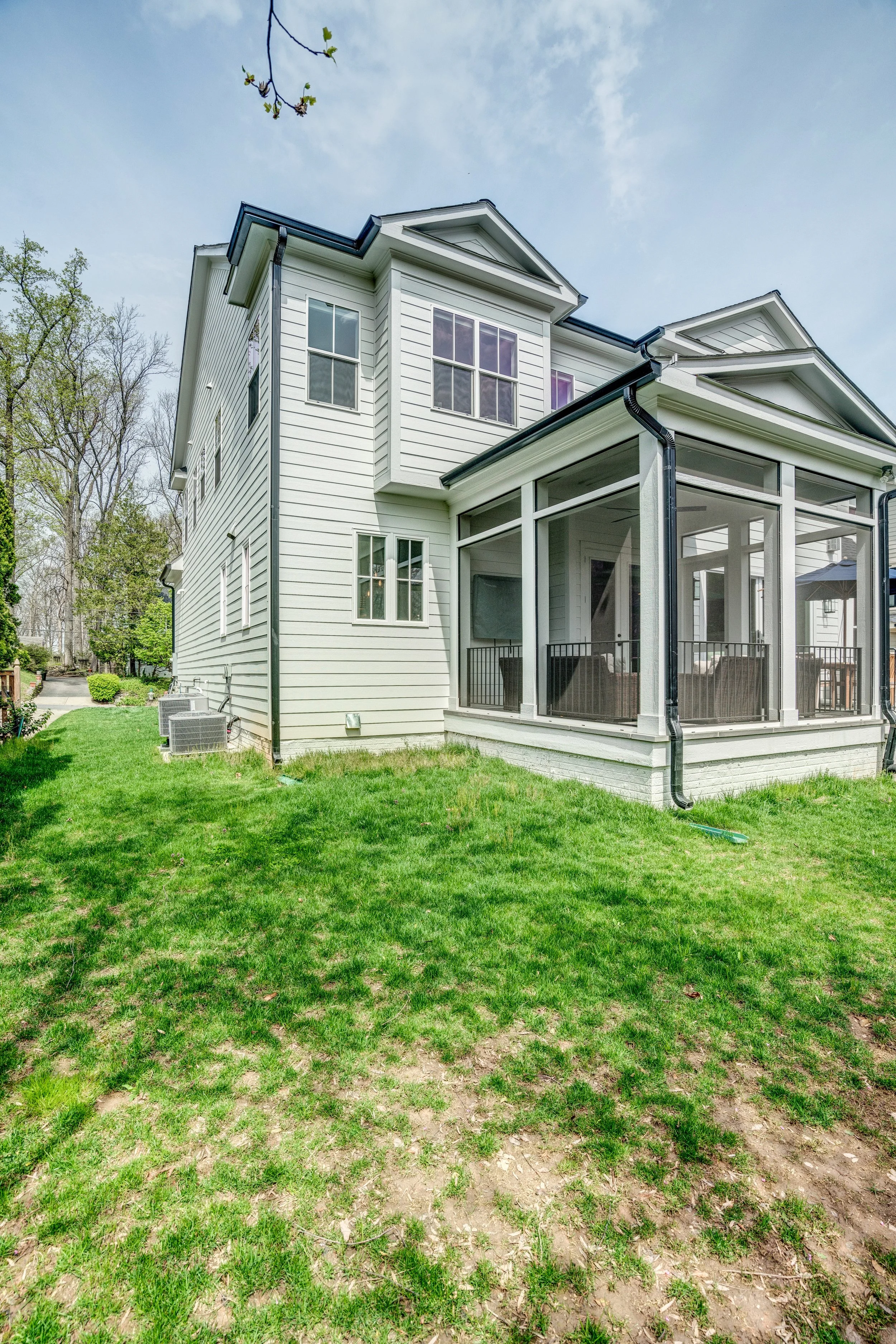 A backyard with a well-maintained lawn, green grass, and some patches of dirt, in front of a white multi-story house with a screened porch and large windows. There are trees along the side of the yard and a cloudy sky overhead.