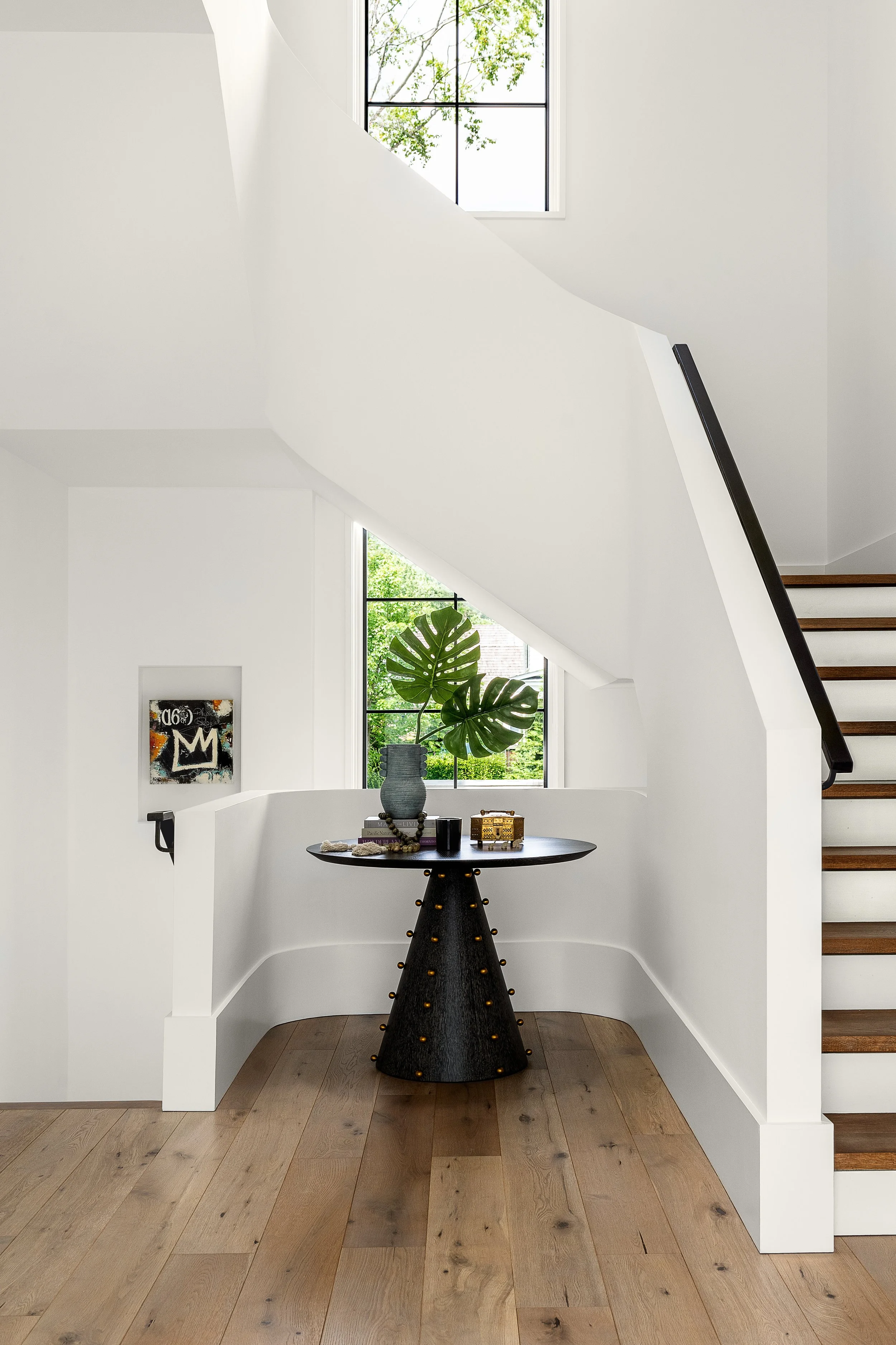 Interior view of a modern, minimalist staircase landing with natural light from large windows, displaying a black round table with decorative items and a large potted plant, light hardwood floors, white walls, and staircase with wooden steps and blac
