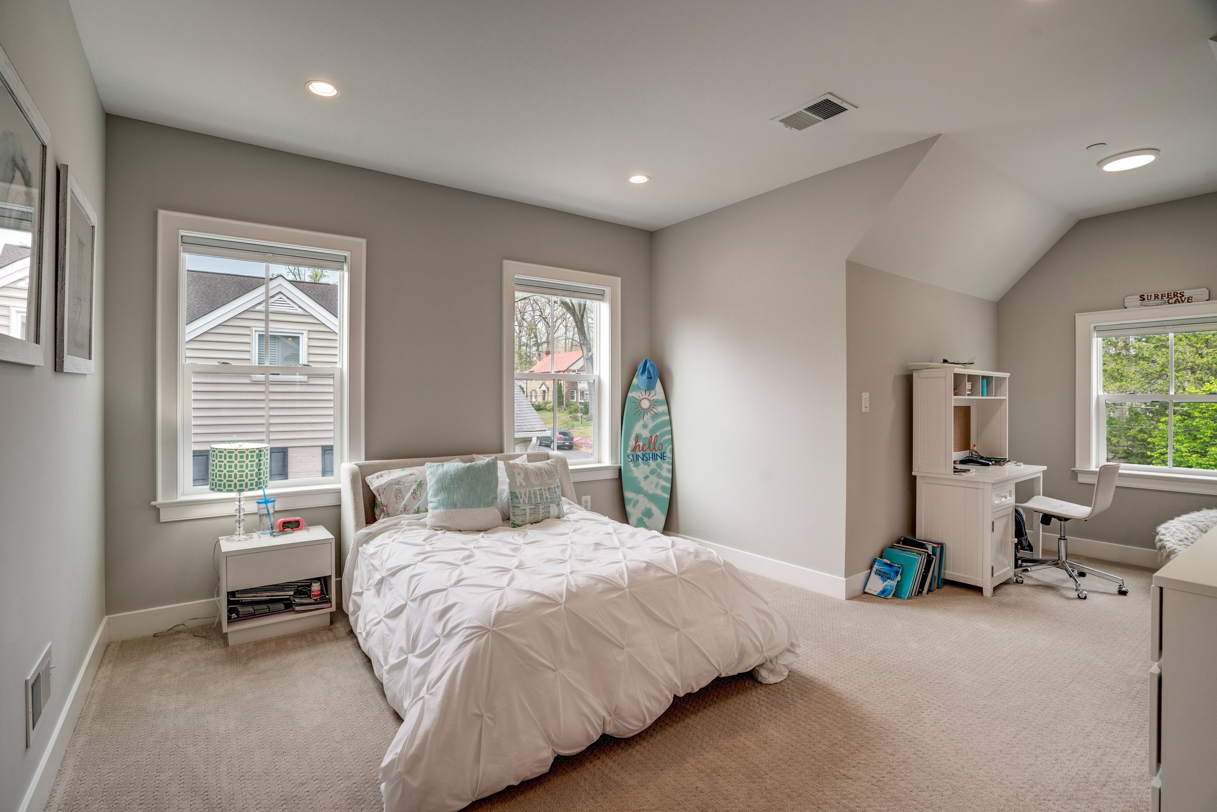 A bedroom with two large windows, a bed with white bedding and pillows, a white nightstand with a lamp and items, a surfboard with the words 'Hello Sunshine,' a white desk with books, an office chair, and framed pictures on the wall.