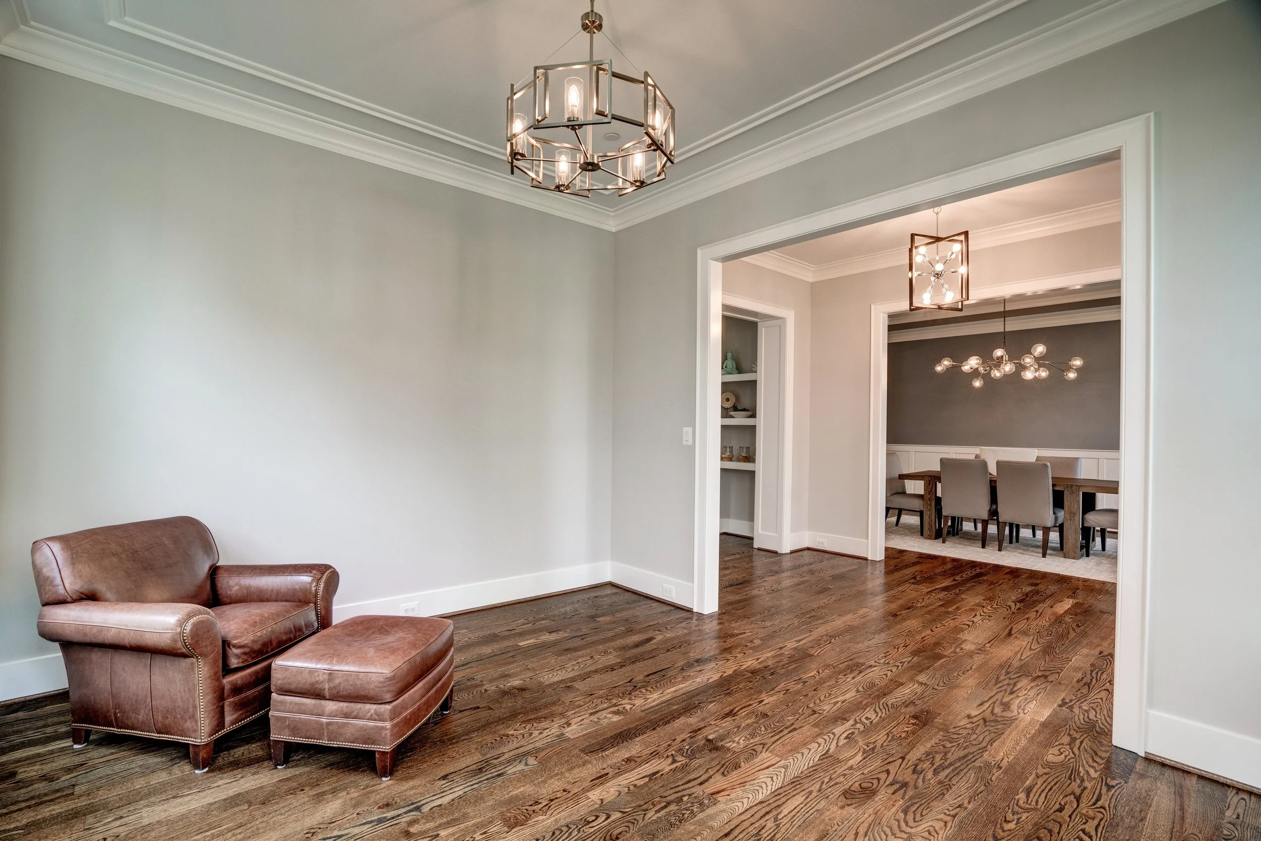 Living room with brown leather armchair and ottoman, hardwood floors, light green walls, and two modern chandeliers in adjacent rooms.