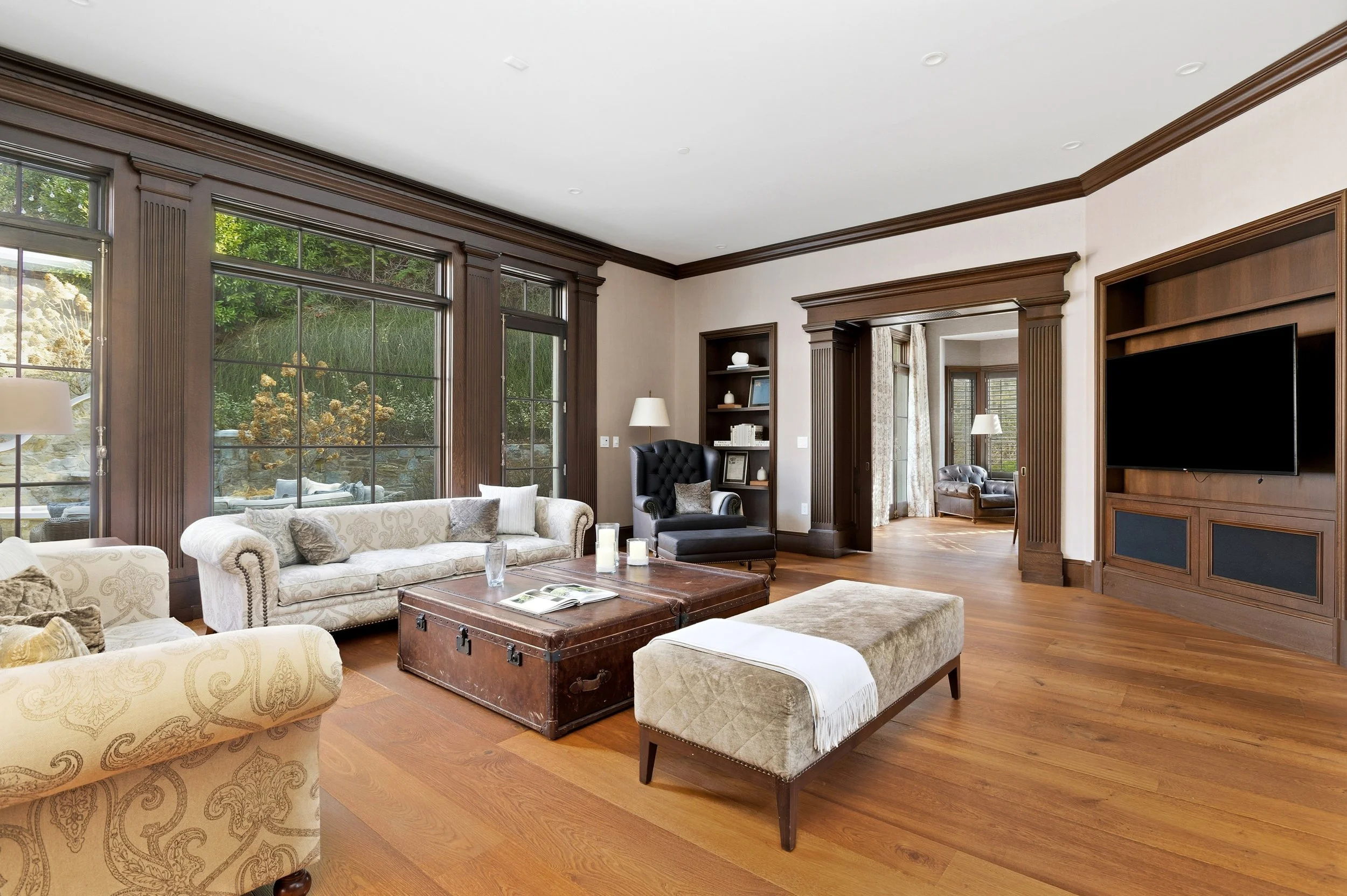 Living room with large windows, beige and black armchairs, a wooden coffee table, a built-in wooden entertainment center, and hardwood flooring.