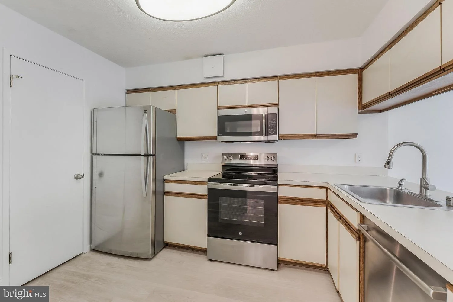 Kitchen with beige cabinets, stainless steel refrigerator, microwave, oven, and sink, on light-colored flooring and white walls.