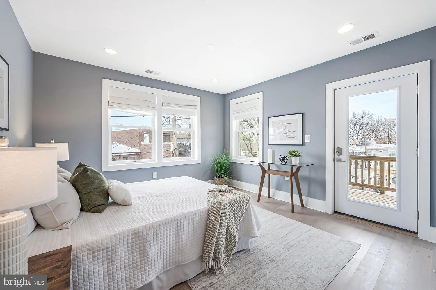 Bright bedroom with a white bed, green pillow, and beige blanket, gray walls, wooden floor, large windows, and glass door leading to a balcony with outdoor view.