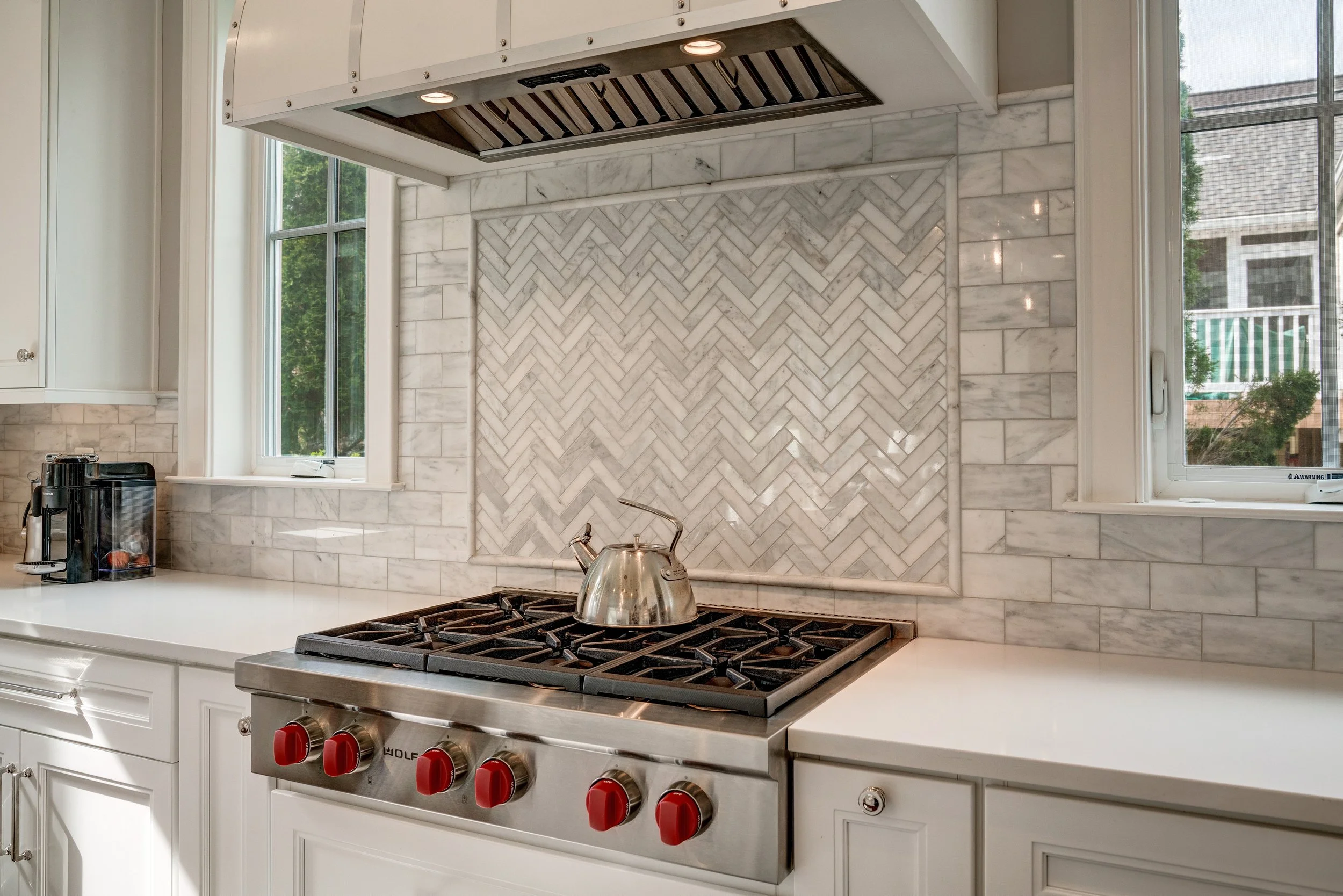 Kitchen with white cabinets, marble backsplash in herringbone pattern, stainless steel gas stove with red knobs, and windows showing greenery outside.