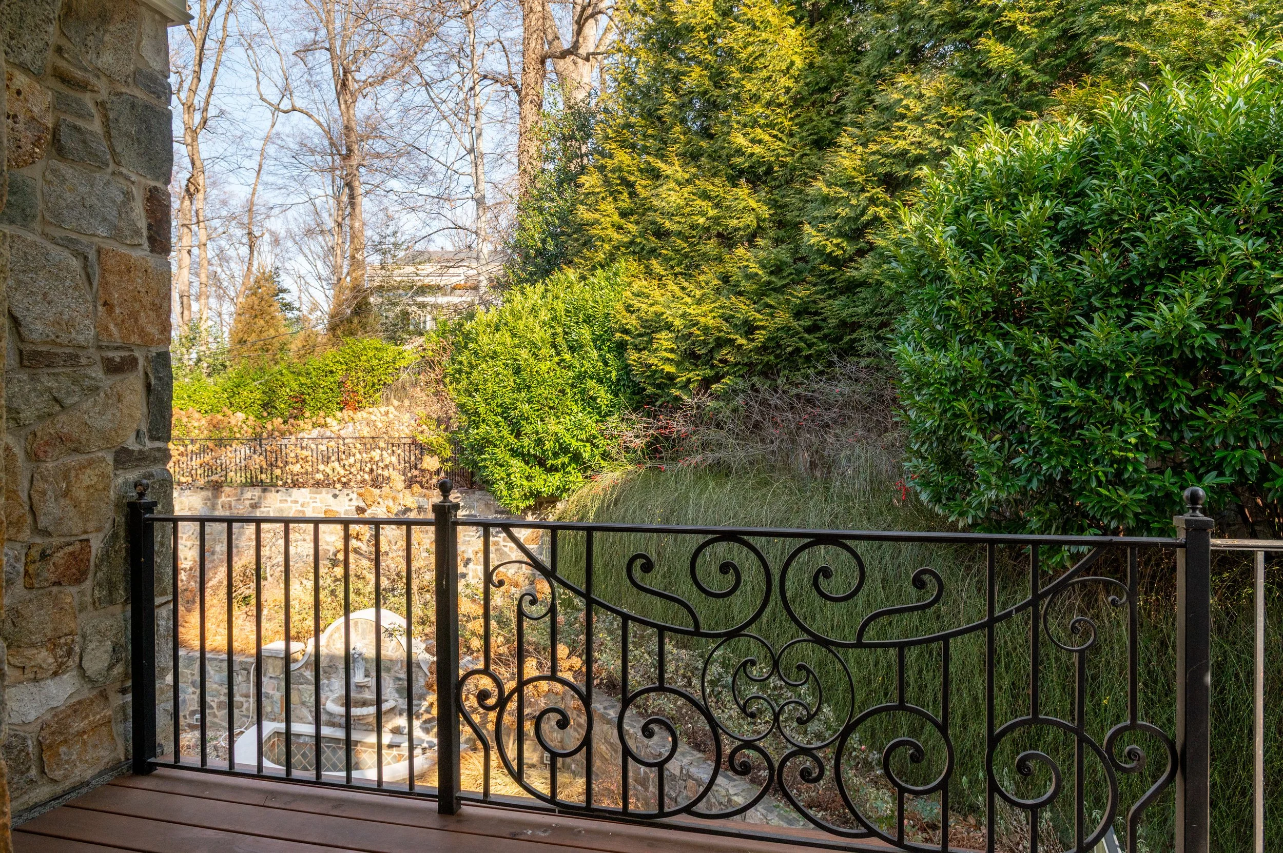 View from a balcony with a decorative metal railing, overlooking a courtyard with stone walls, stairs, and lush green trees and bushes.
