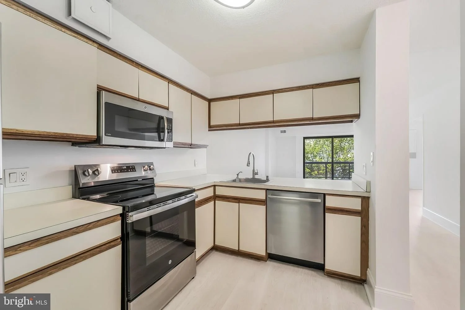 Kitchen with cream cabinets, wooden trim, stainless steel stove, microwave, sink, and dishwasher, with a window showing trees outside.