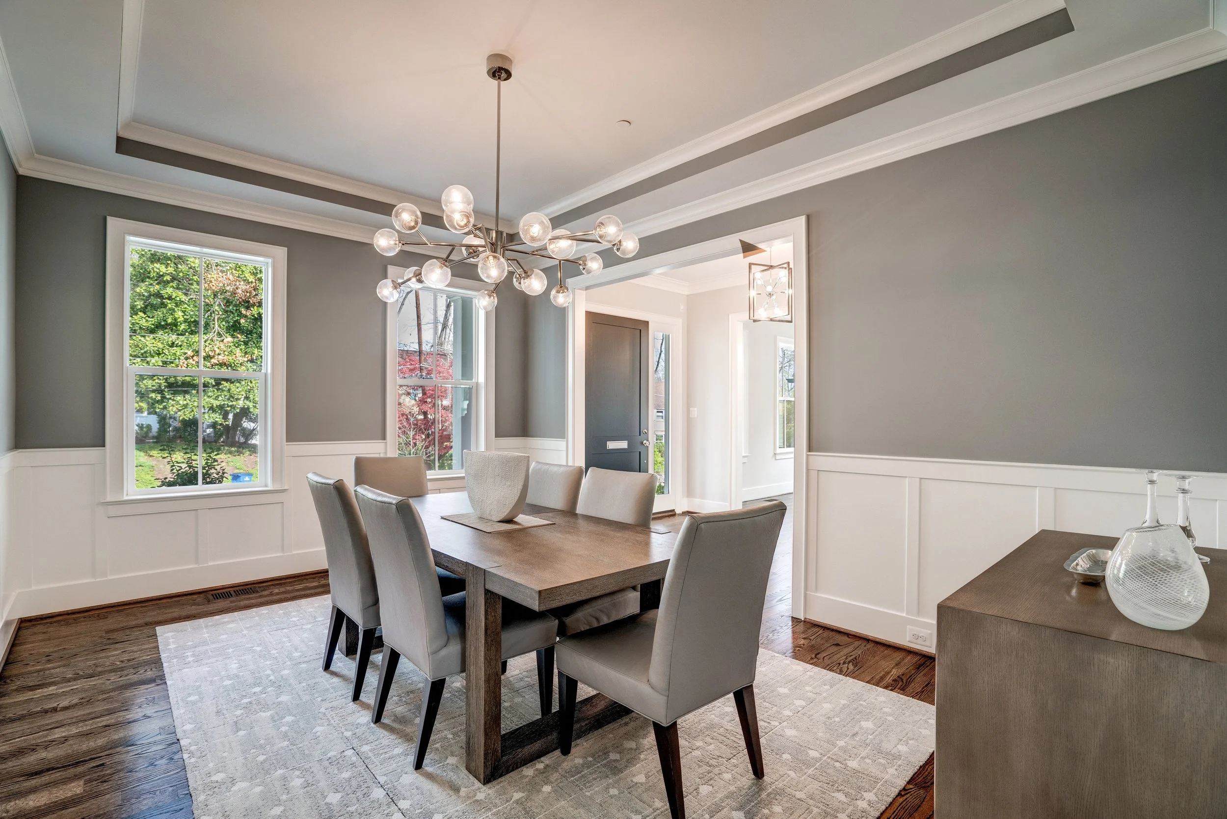 Dining area with a wooden table, beige chairs, large windows showing greenery outside, and a modern chandelier on the ceiling.