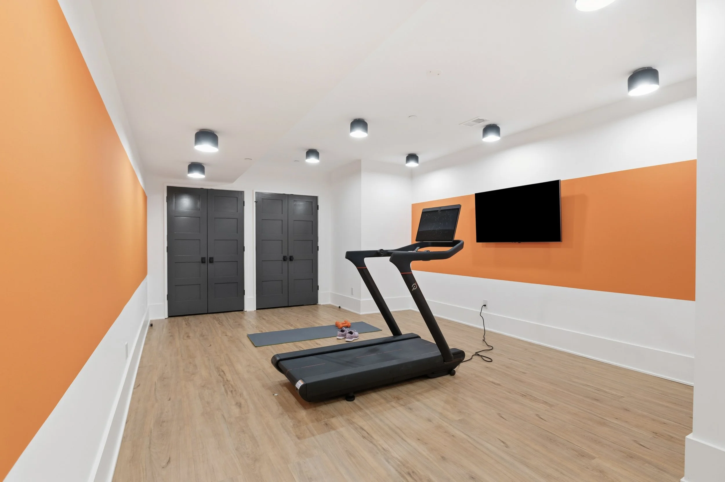 A home gym with orange accent walls, a treadmill, a workout mat, shoes, orange dumbbells, a mounted flat-screen TV, and gray doors.