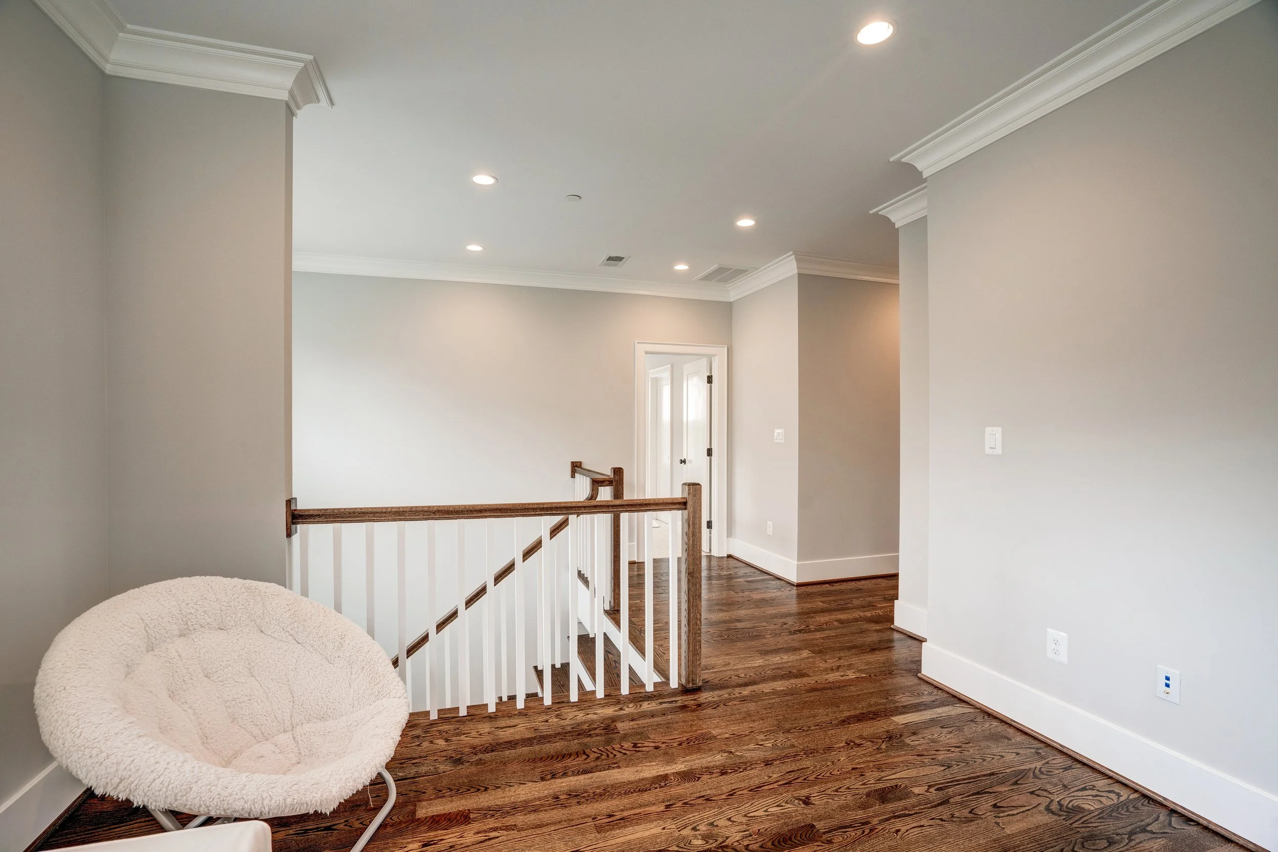 Interior view of a modern upstairs area with hardwood floors, light-colored walls, a white rail with wooden handrails, a door, and recessed ceiling lights. A beige plush chair is partially visible in the foreground.