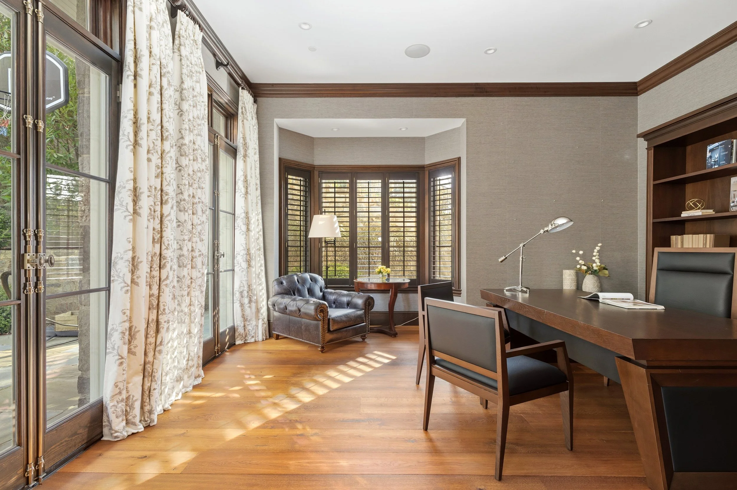A bright living room with large windows, wooden floors, a leather armchair, a wooden coffee table with a small vase of yellow flowers, a floor lamp, and built-in bookshelves.
