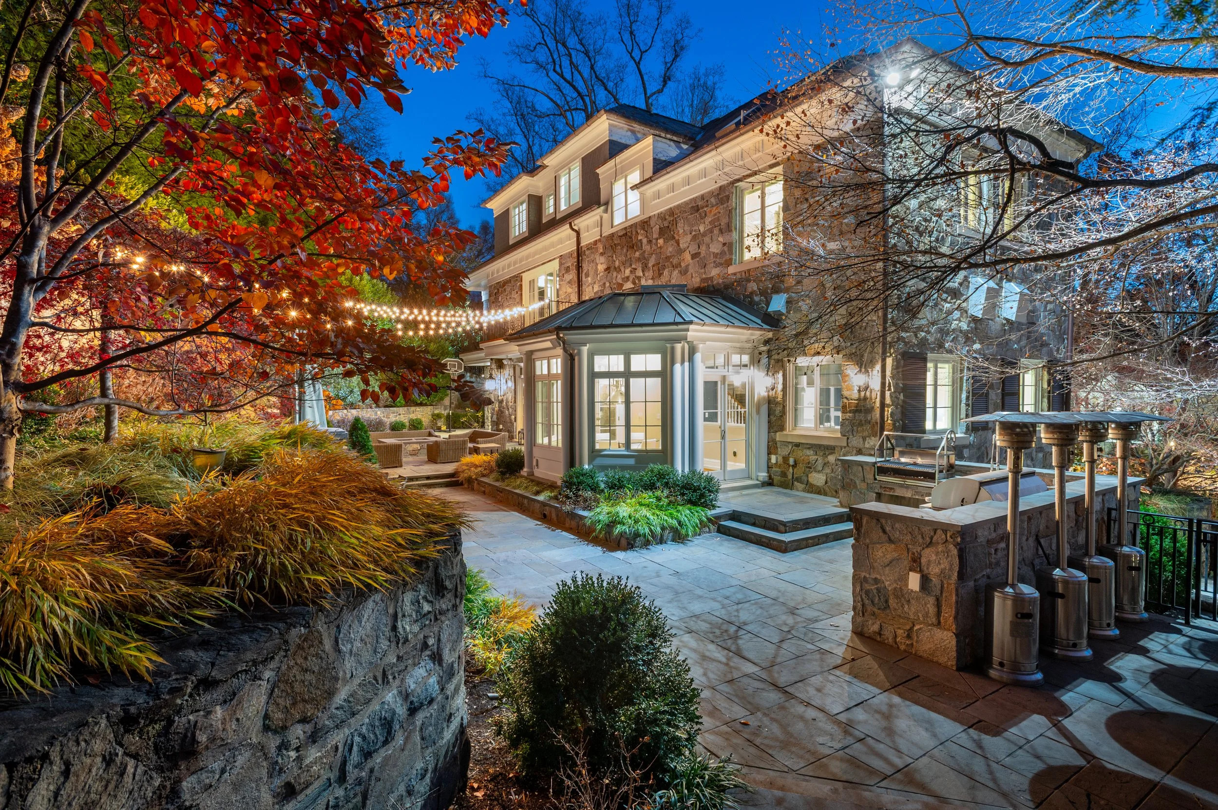 A large house surrounded by trees with autumn leaves, illuminated by outdoor lighting, with a patio area and a well-maintained garden.