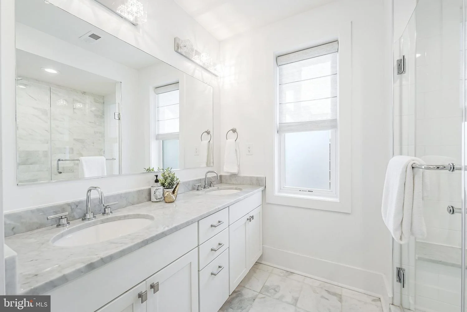 Bright white bathroom with double vanity, marble countertop, large mirror, soap dispenser, potted plant, towel rings with towels, window with white blinds, glass shower enclosure, and tiled floor.