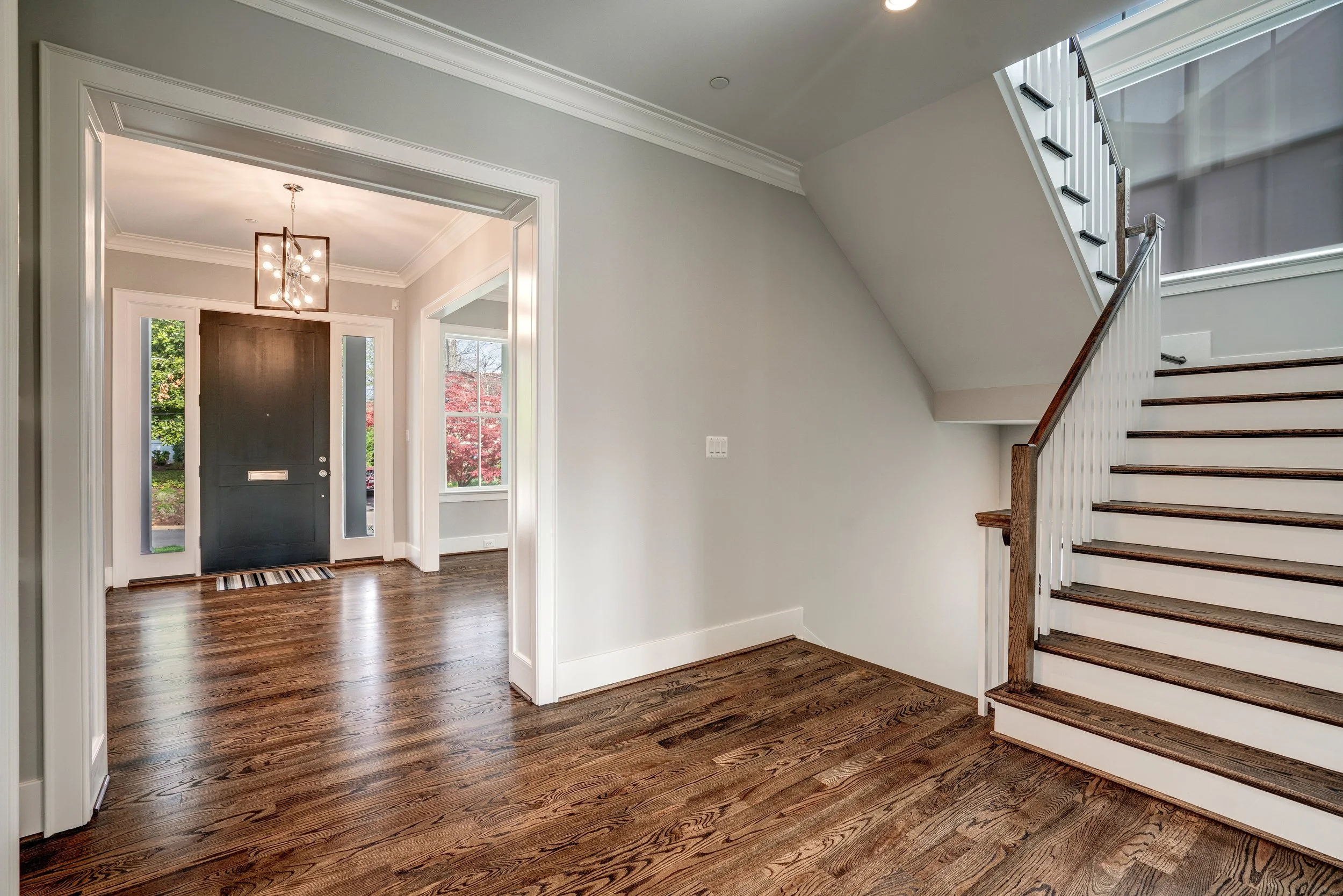 View of a modern home's entrance with hardwood floors, a front door, a staircase with wooden steps and white balusters, and large windows showing outdoor greenery.