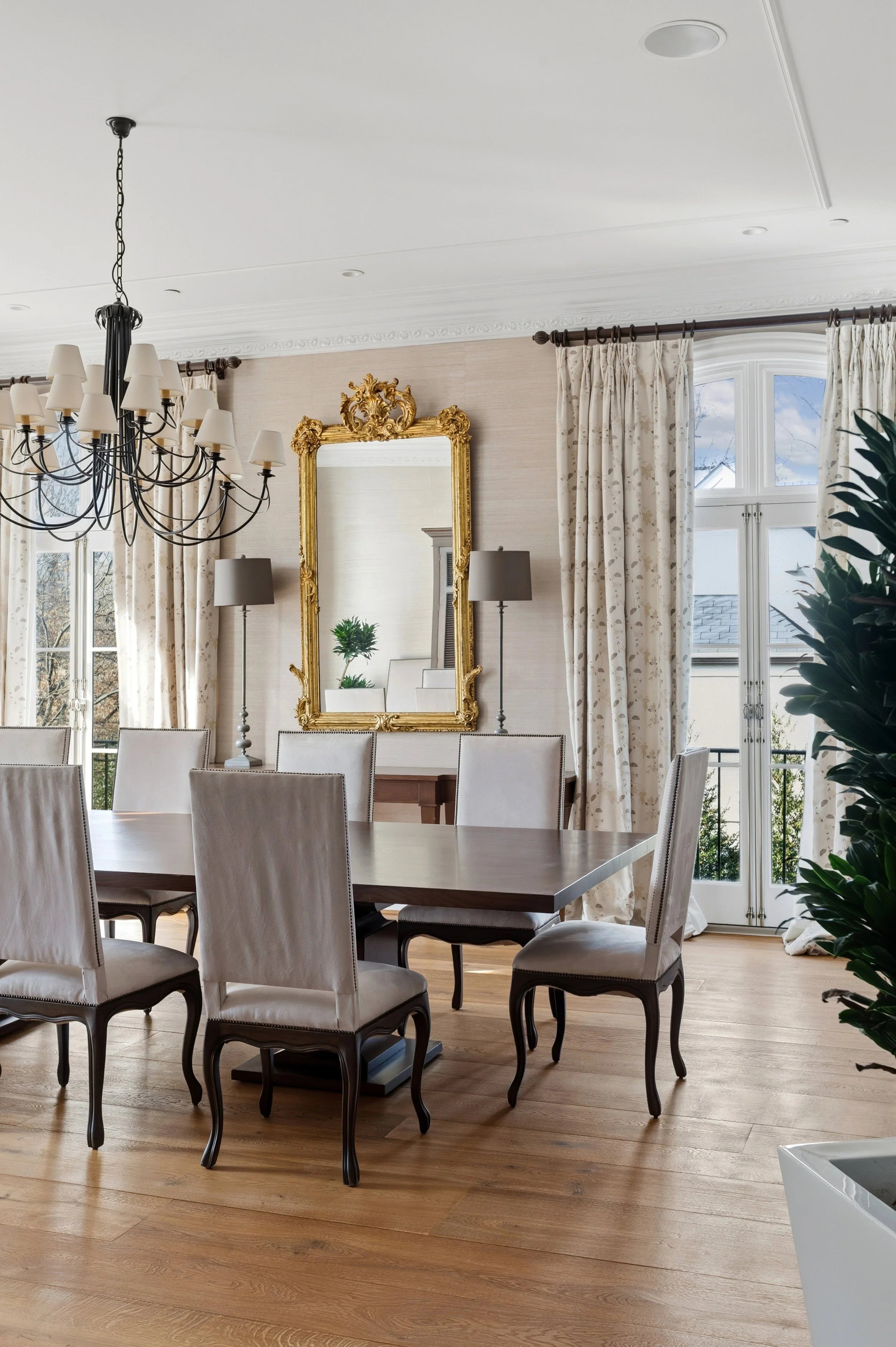 Elegant dining room with a large rectangular wooden table surrounded by cream upholstered chairs, a black wrought-iron chandelier with cream lampshades hanging from the ceiling, a gold ornate mirror on a beige wall, two tall lamps with grey shades, a