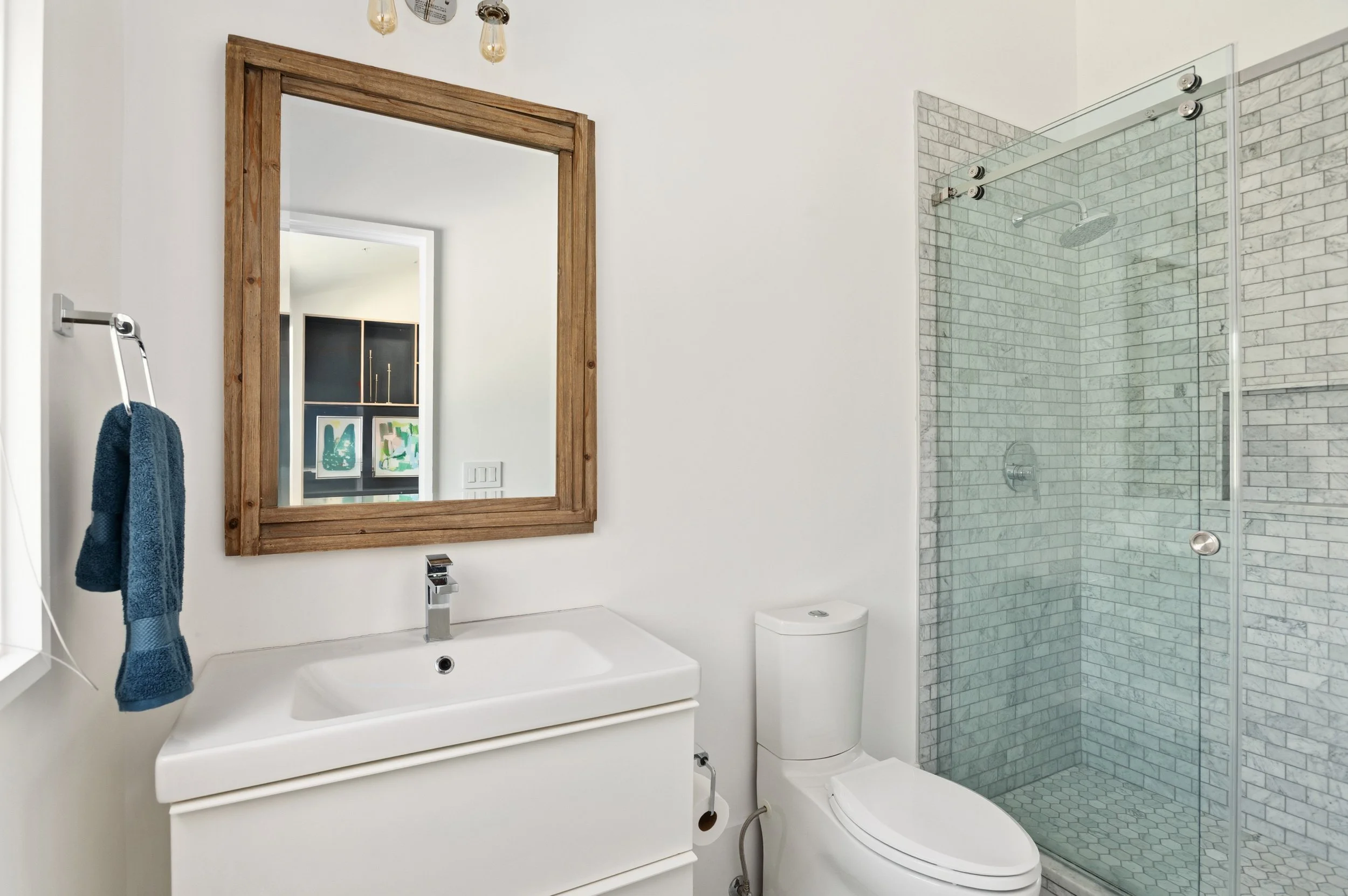 Modern bathroom with a white sink and wooden framed mirror, a floating towel rack with a blue towel, a white toilet, and a glass-enclosed shower with gray tile walls.