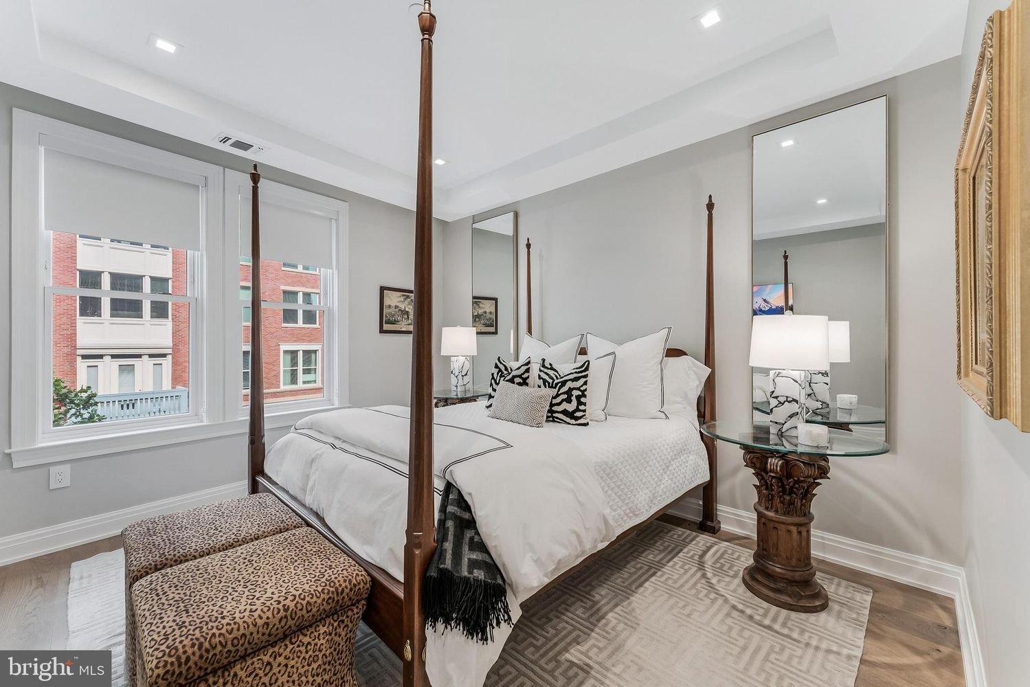 A bedroom with a four-poster bed, white bedding, and black-and-white accent pillows. There are two windows with white blinds, a round glass side table with a lamp, and two framed pictures on the wall. An animal print ottoman at the foot of the bed, a