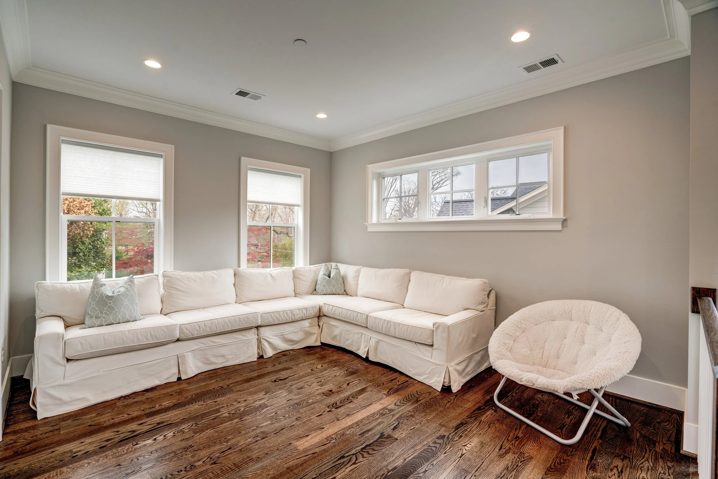 A living room with a large white sectional sofa, two decorative pillows, and a round white pet bed on a wooden floor, with multiple windows bringing in natural light.