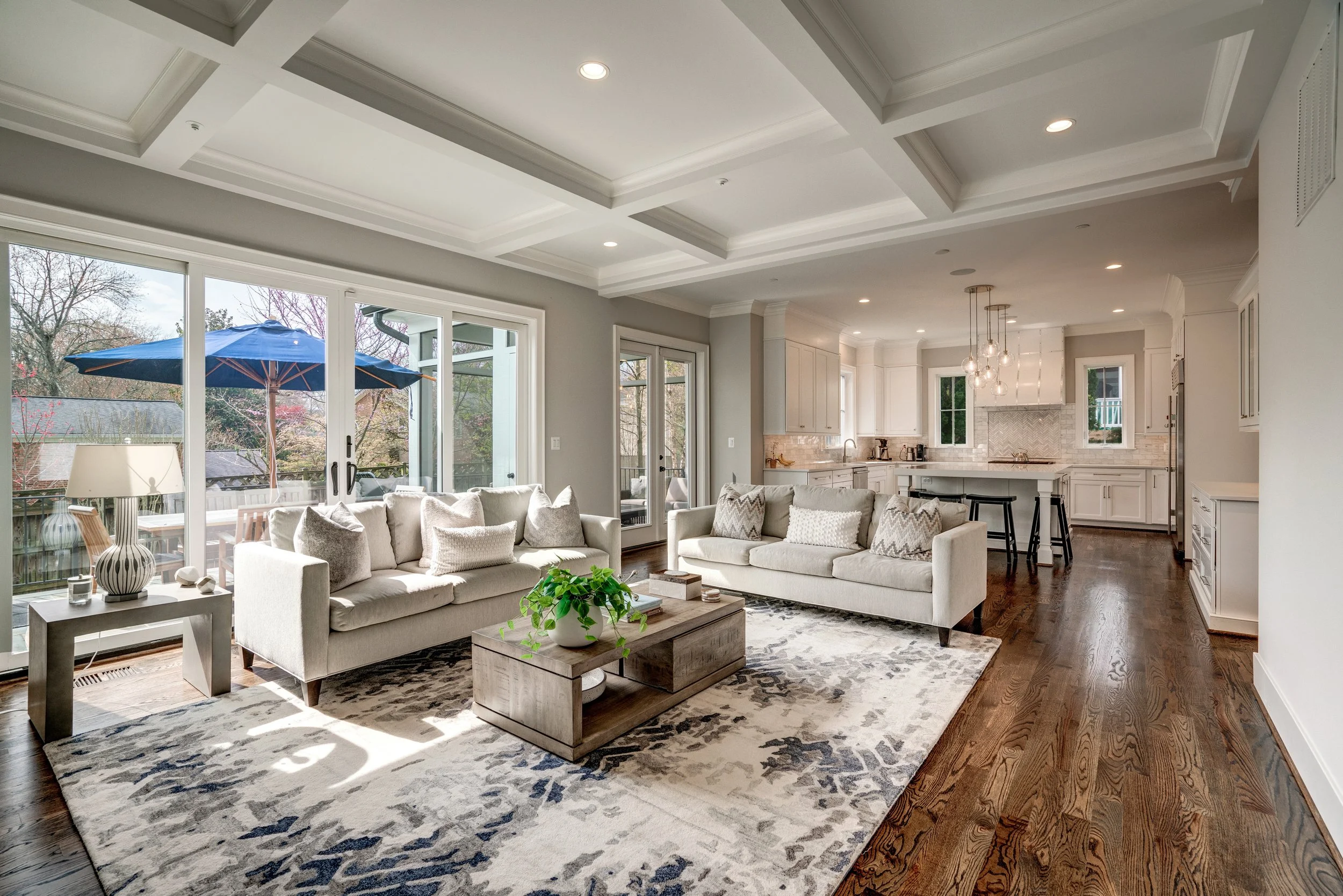 Open-concept living room and kitchen with white furniture, large windows, hardwood floors, and a view of an outdoor patio with a blue umbrella.