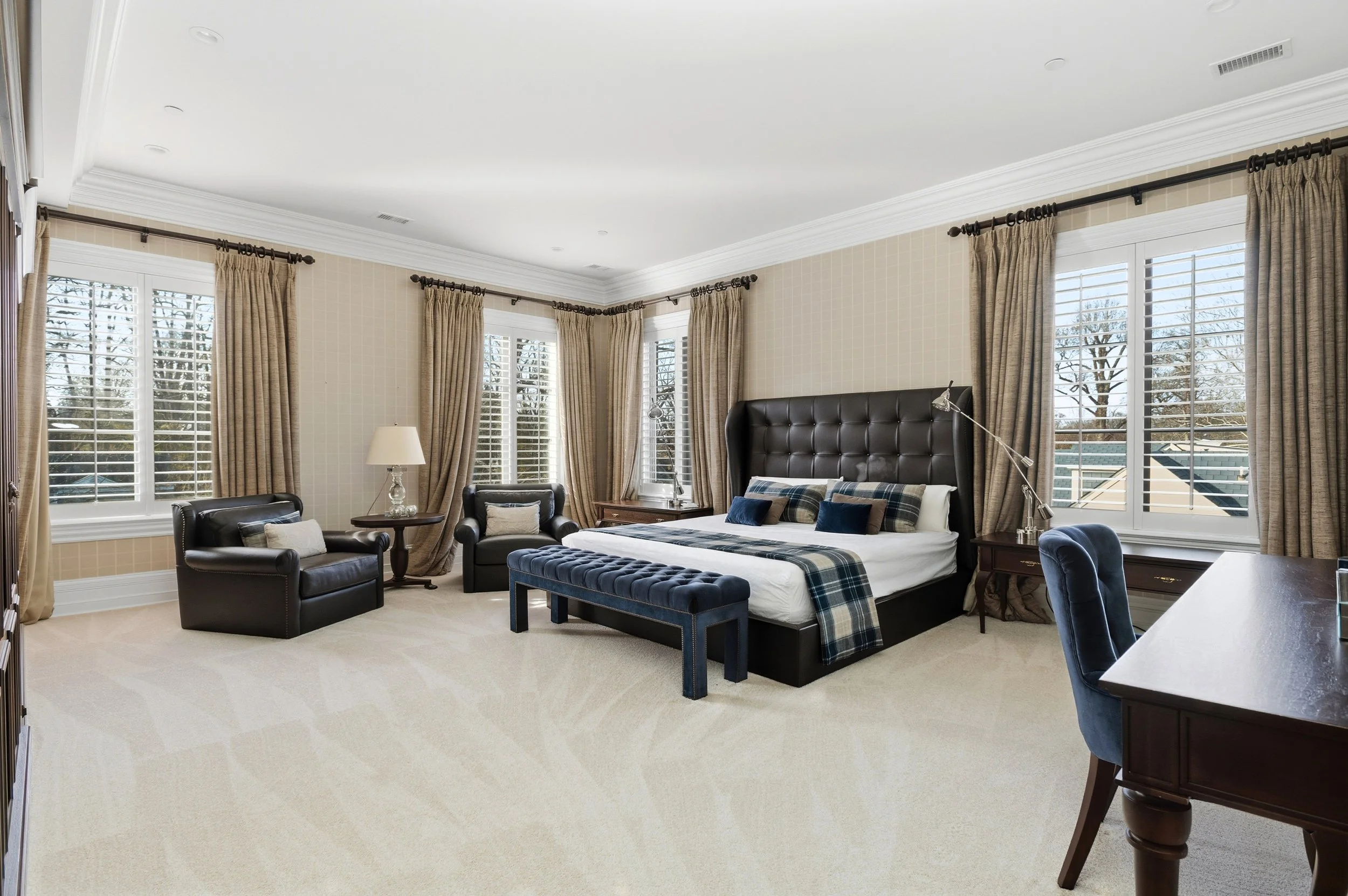 A spacious bedroom with large windows covered by beige curtains, a black tufted bed with plaid bedding, two black chairs with pillows, a side table with a lamp, and a desk with a blue upholstered chair. Outside the windows, snow-covered trees are vis
