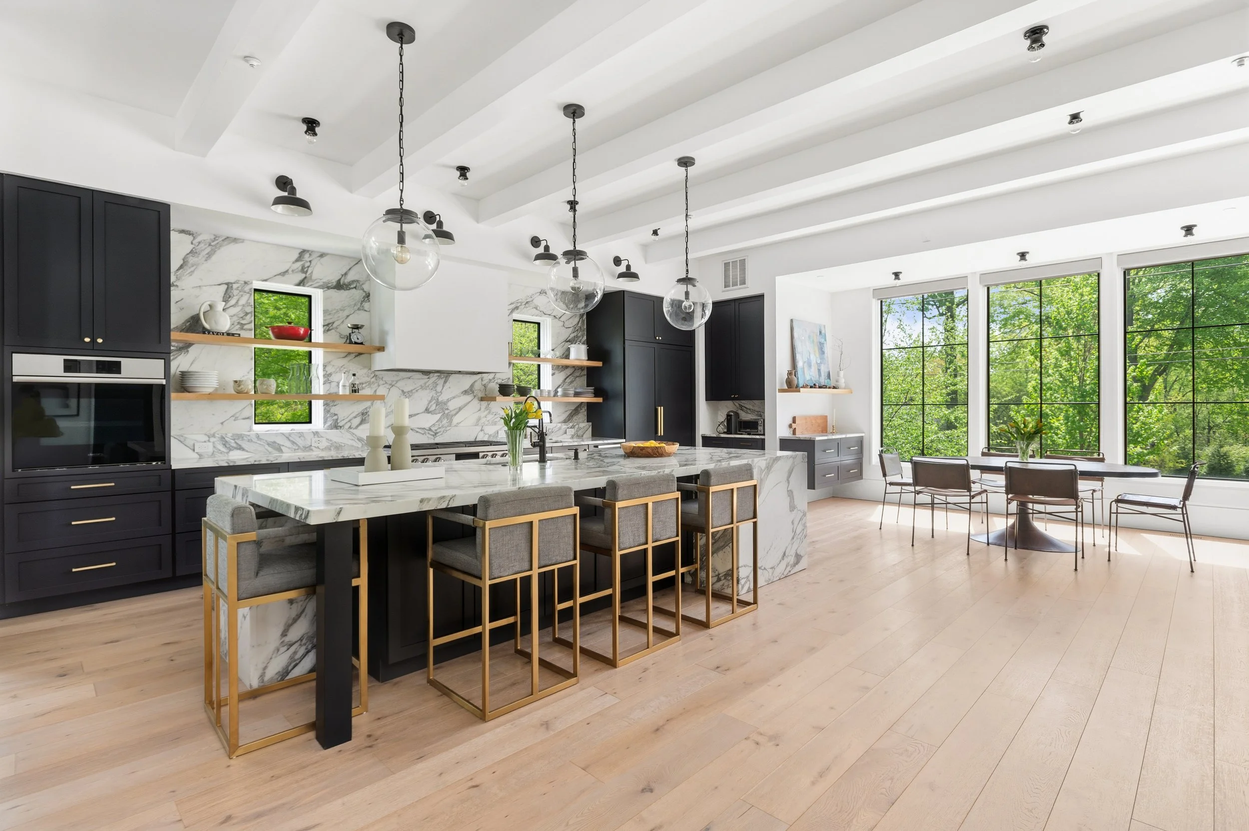 Modern kitchen with black cabinets, marble island, open shelving, large windows with greenery outside, and a dining area with a round table and chairs.