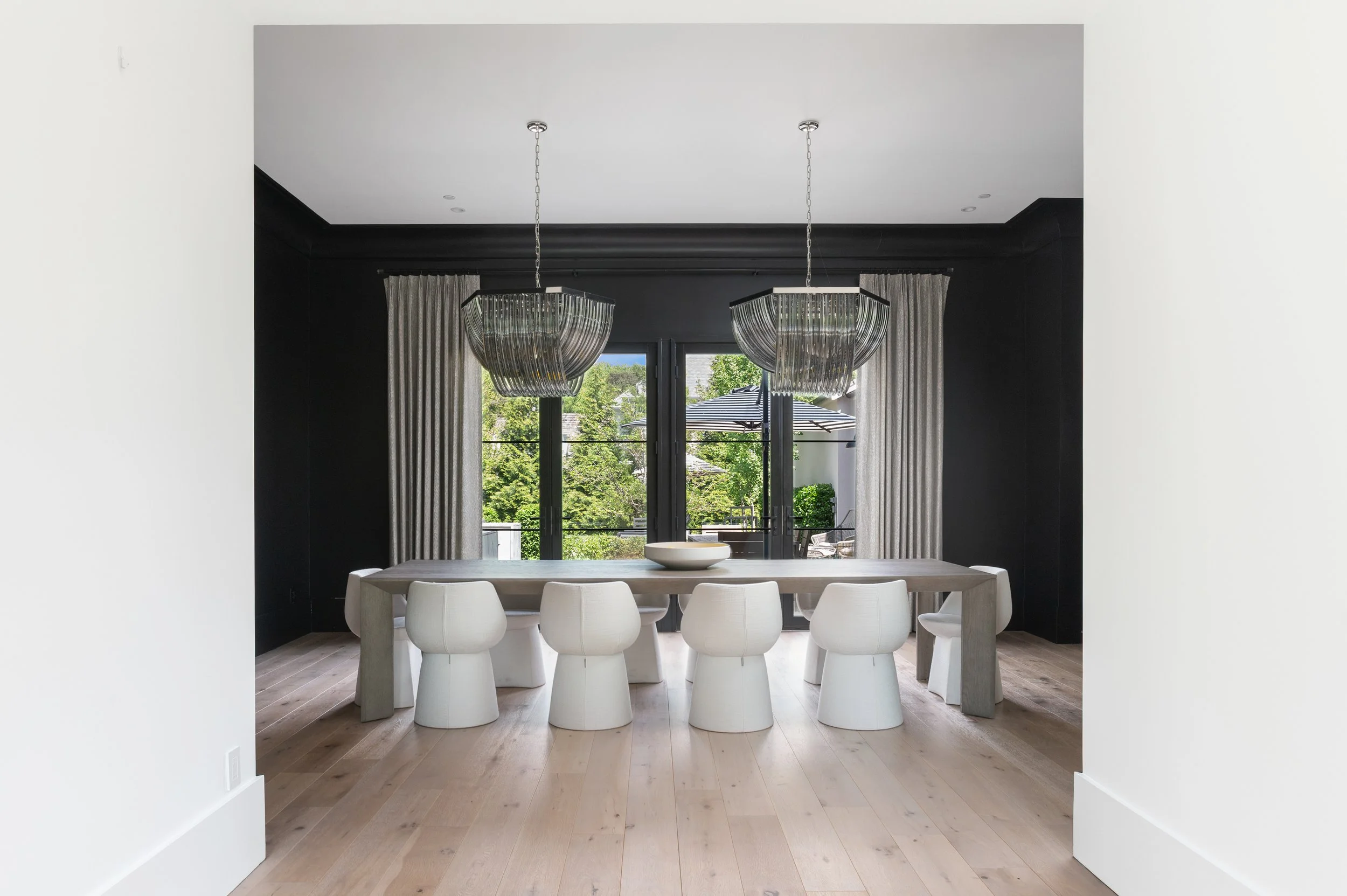 Modern dining room with white oval table, white chairs, black walls, large windows with curtains, and two hanging glass light fixtures.