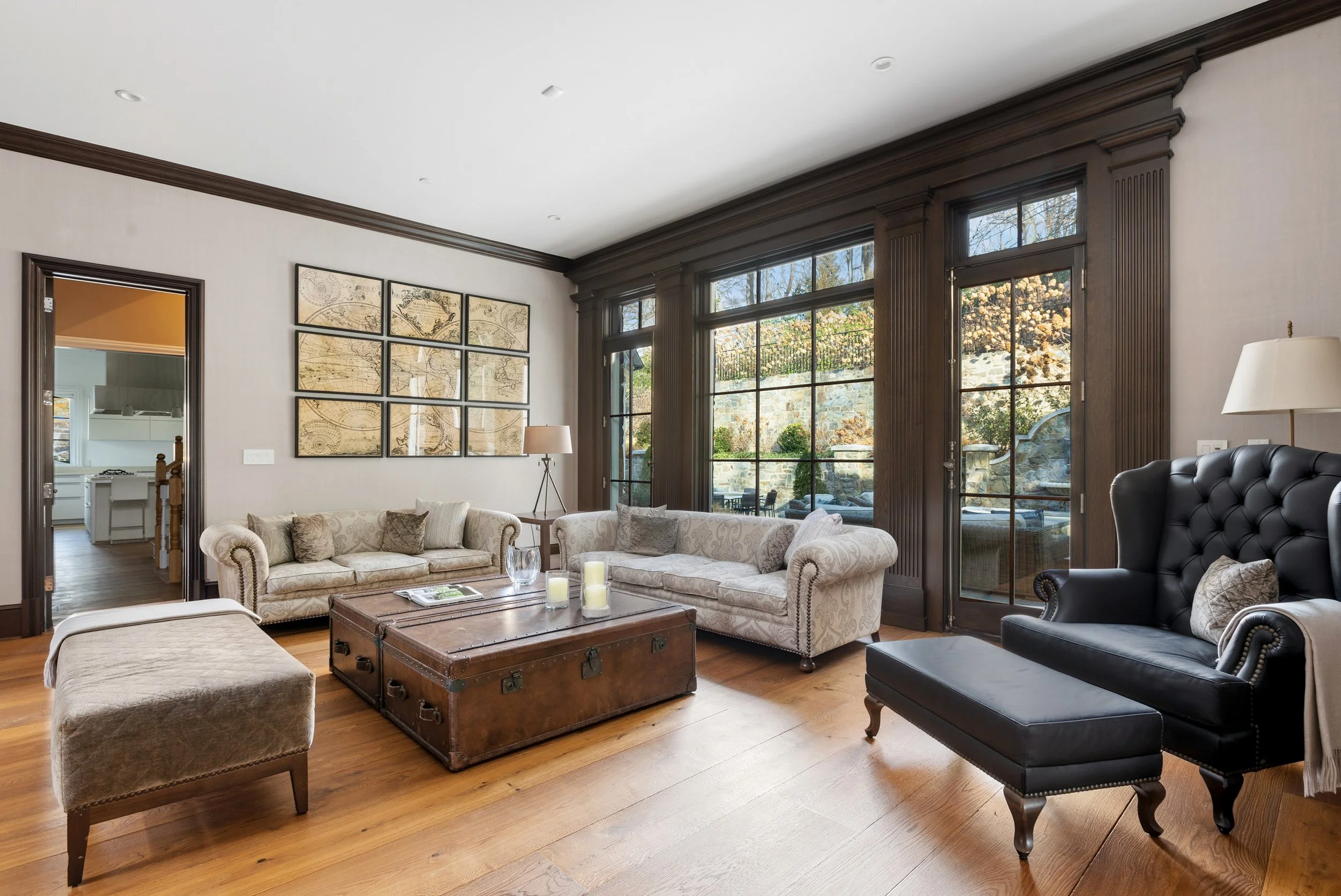 Living room with large window, beige couches, black leather armchair, wooden coffee table with candles, framed map art, and hardwood floors